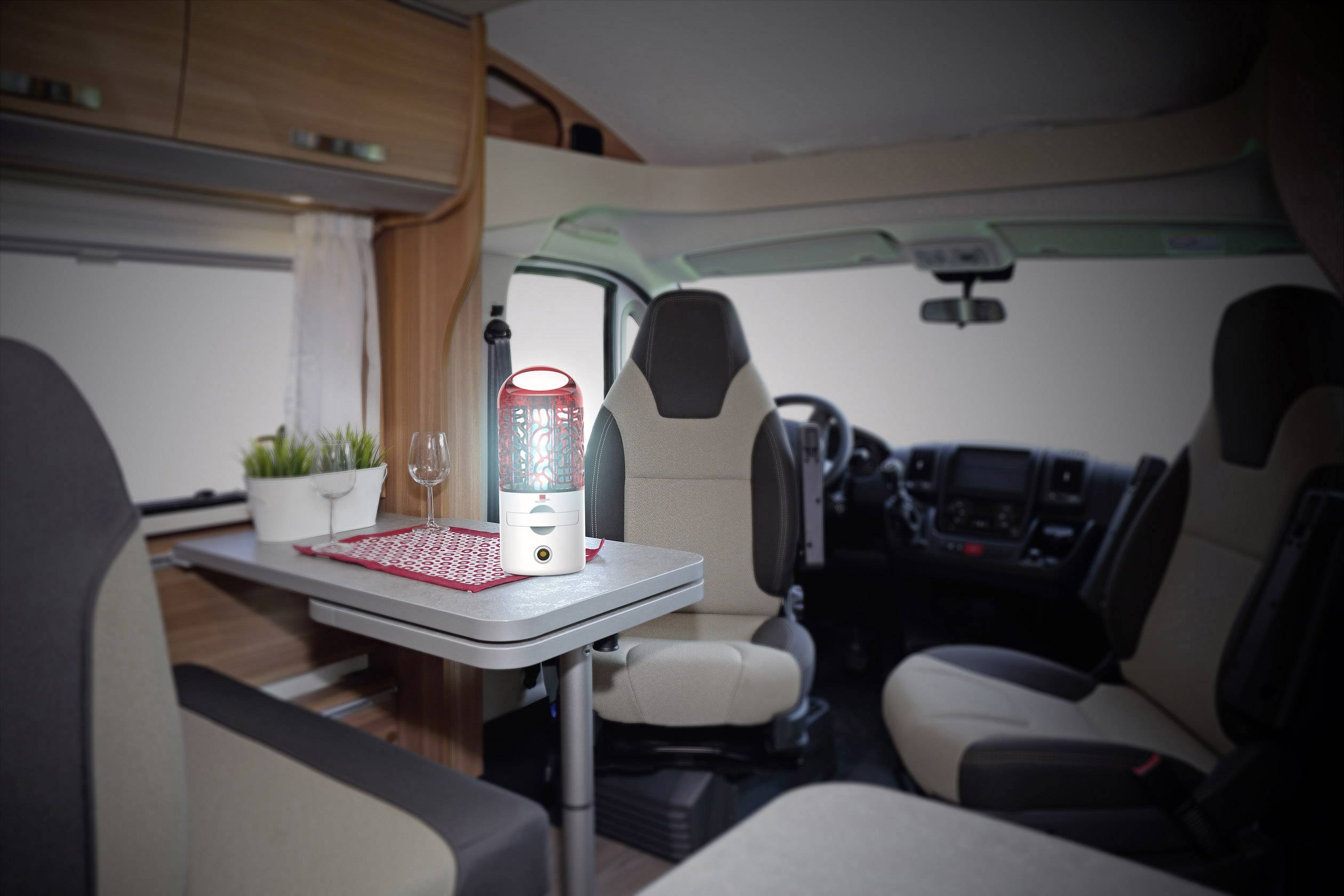 Innenansicht eines Wohnmobils mit Tisch und zwei Sitzen, dekoriert mit einem Topfpflanzen, einem Glas und einer roten Laterne auf dem Tisch.