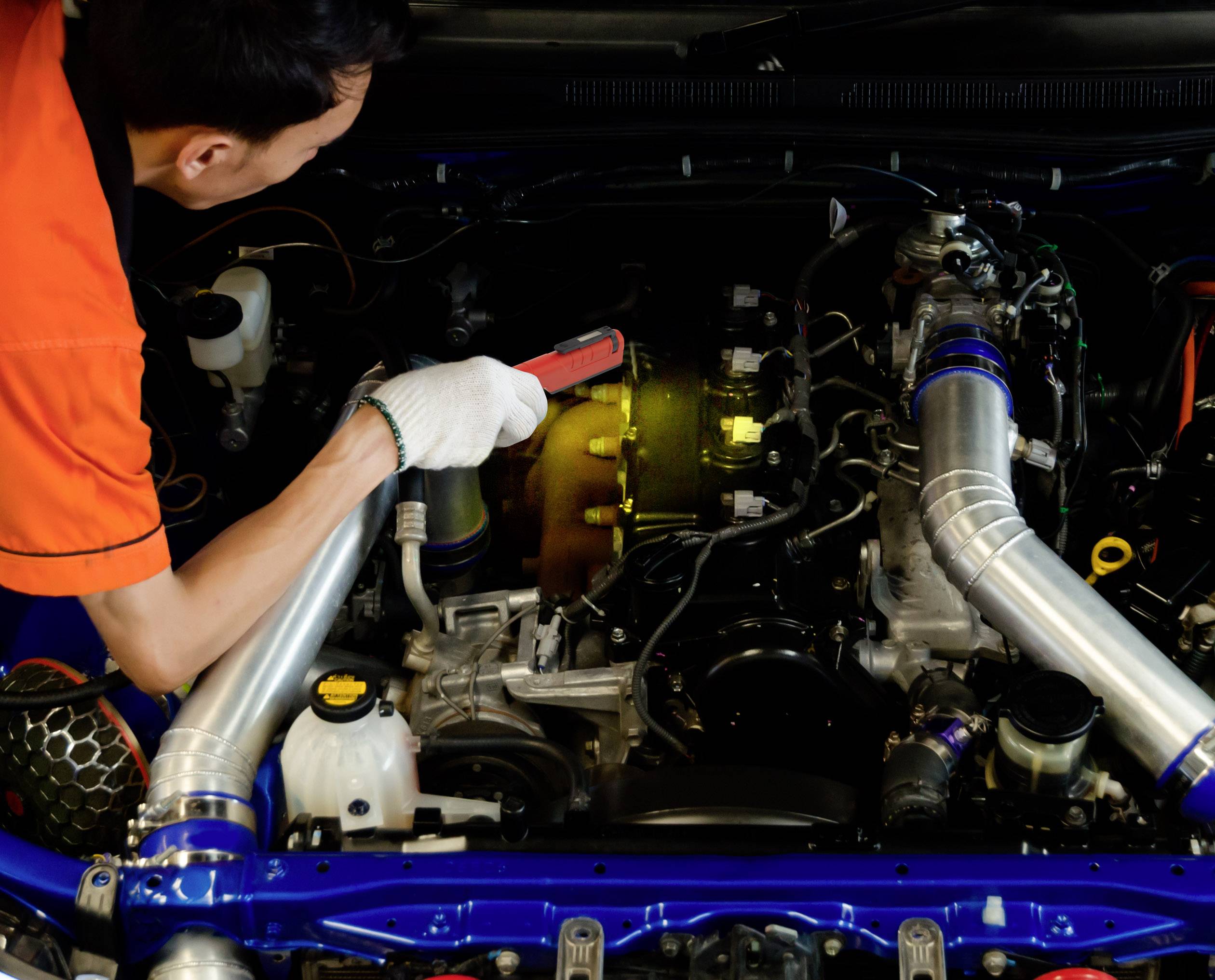 Eine Person in orange Kleidung inspiziert den Motor eines Autos mit einer Taschenlampe, Fokus auf technische Details des Motors.