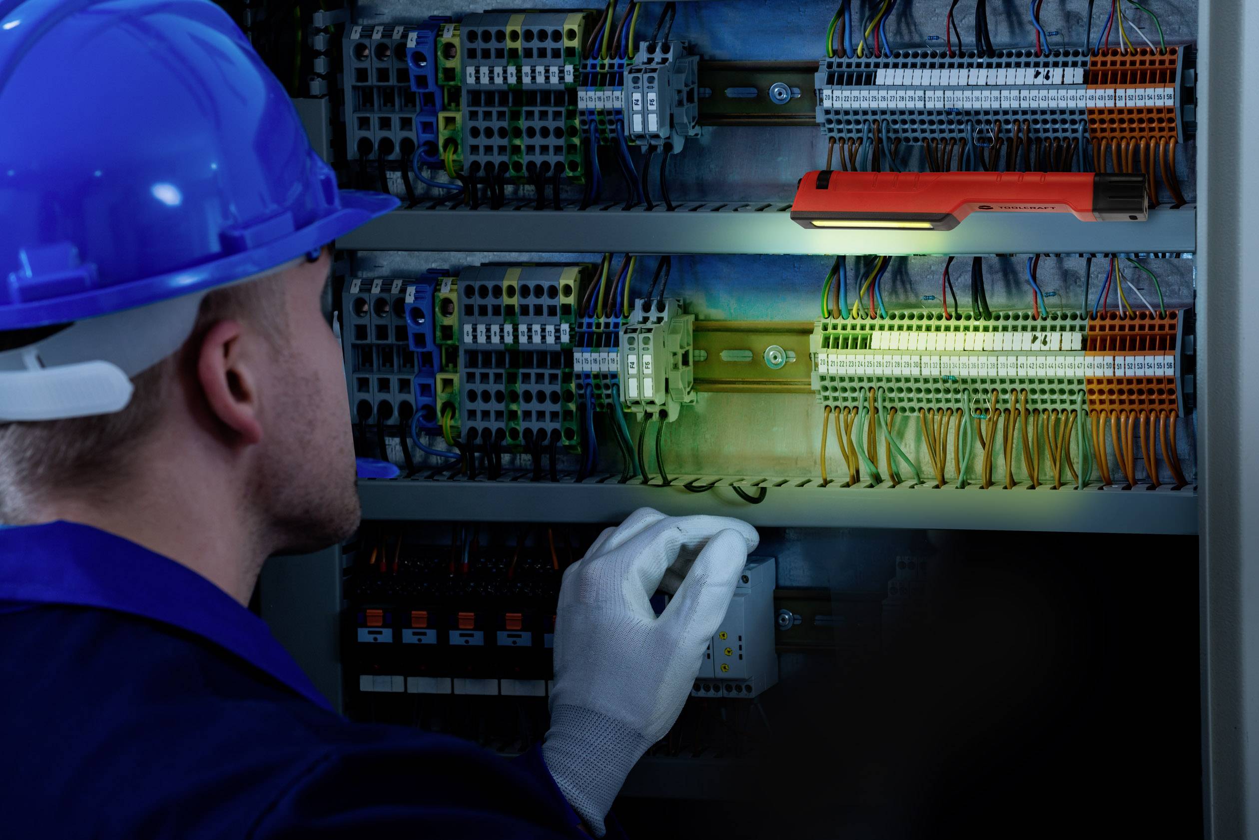 Eine Person in Schutzkleidung überprüft verdrahtete Schaltkästen mit einer Taschenlampe, möglicherweise bei einer elektrischen Inspektion.