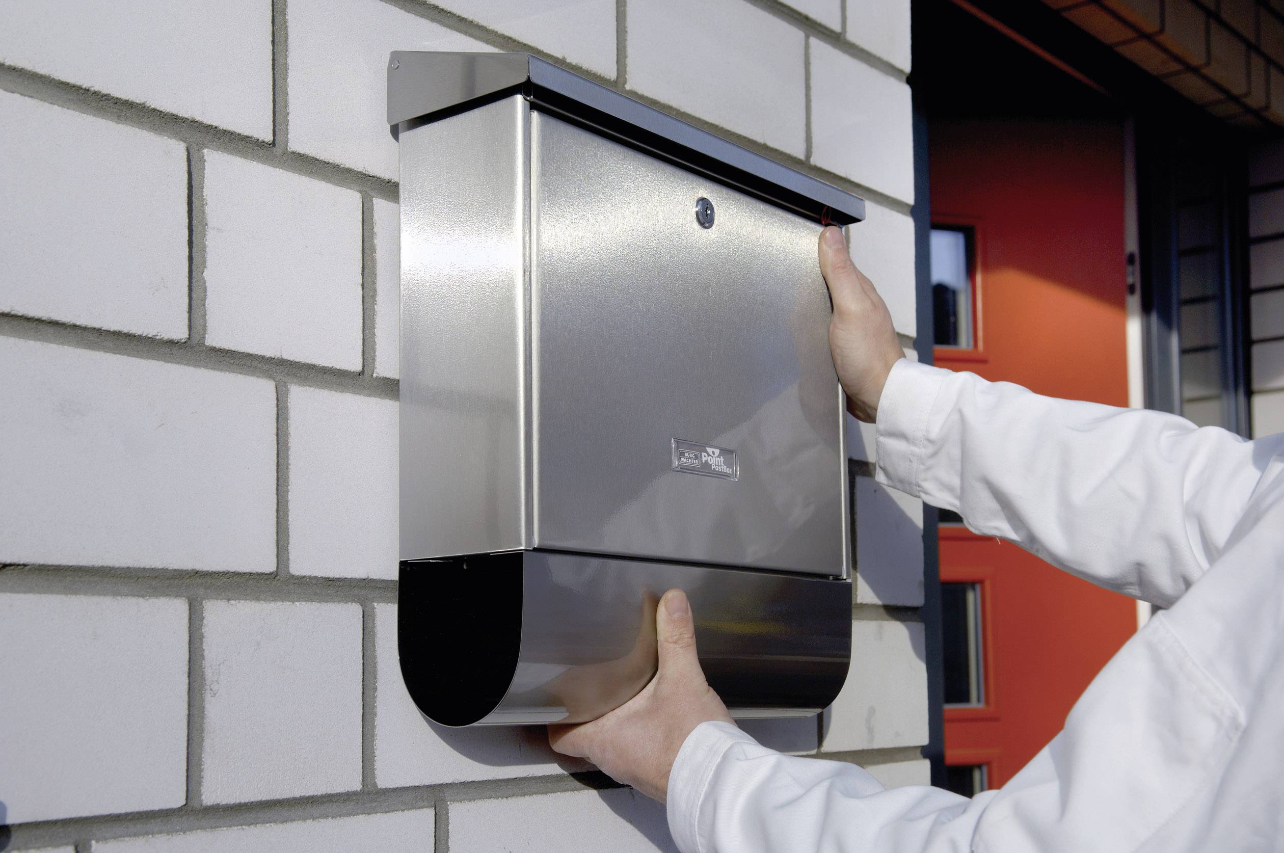 Eine Person installiert einen silbernen Briefkasten aus Metall an einer weißen Ziegelwand. Im Hintergrund ist eine rote Tür zu sehen.