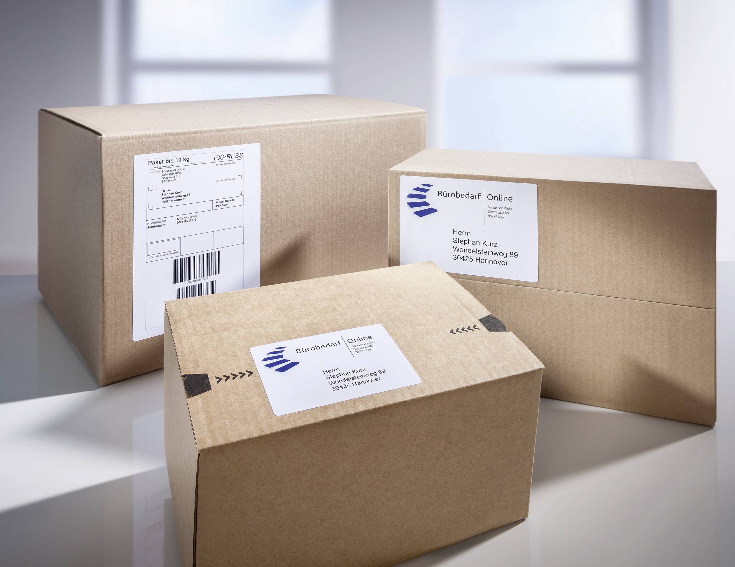 Drei Pakete mit Adressetiketten, die auf einem Tisch stehen. Das mittlere Paket trägt die Aufschrift 'Bürobedarf Online'.
