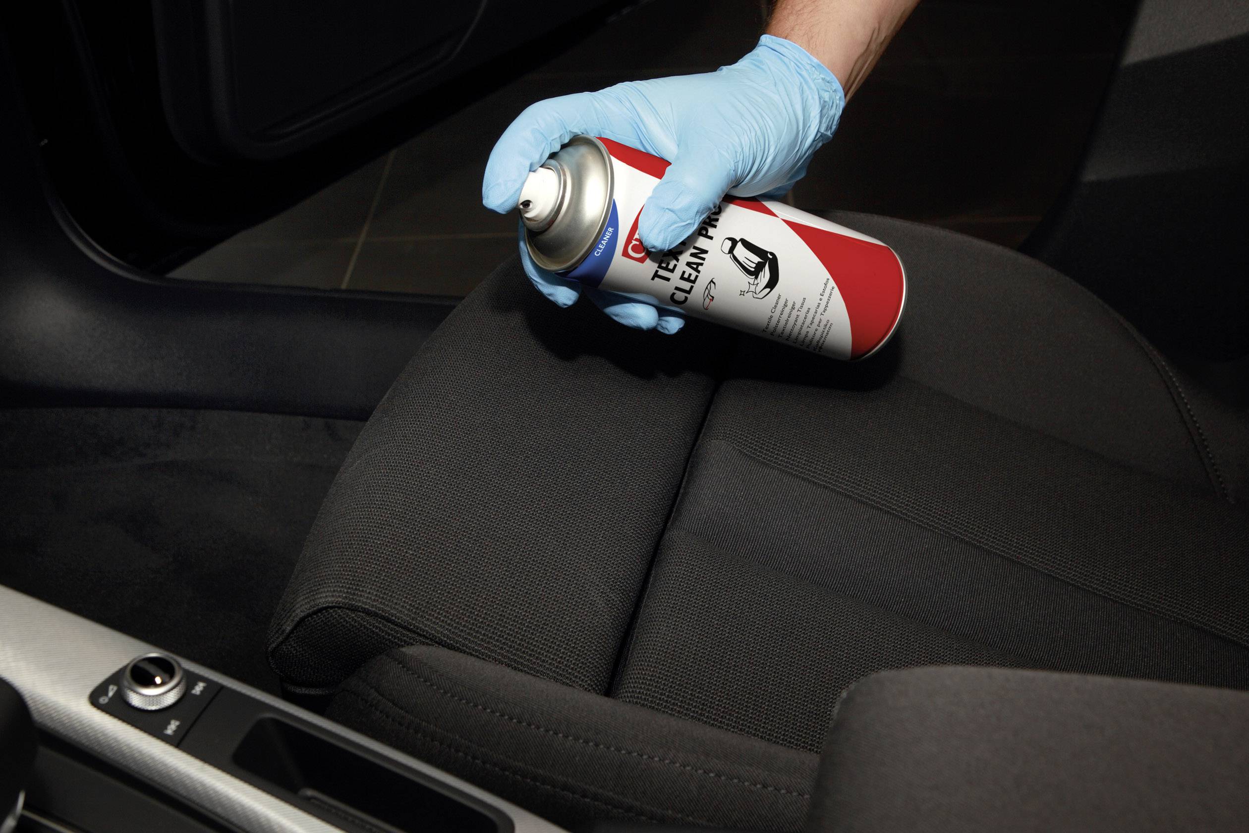 Eine Person mit Handschuhen sprüht Reiniger aus einer Dose auf einen Autositz, um ihn zu reinigen oder zu pflegen.