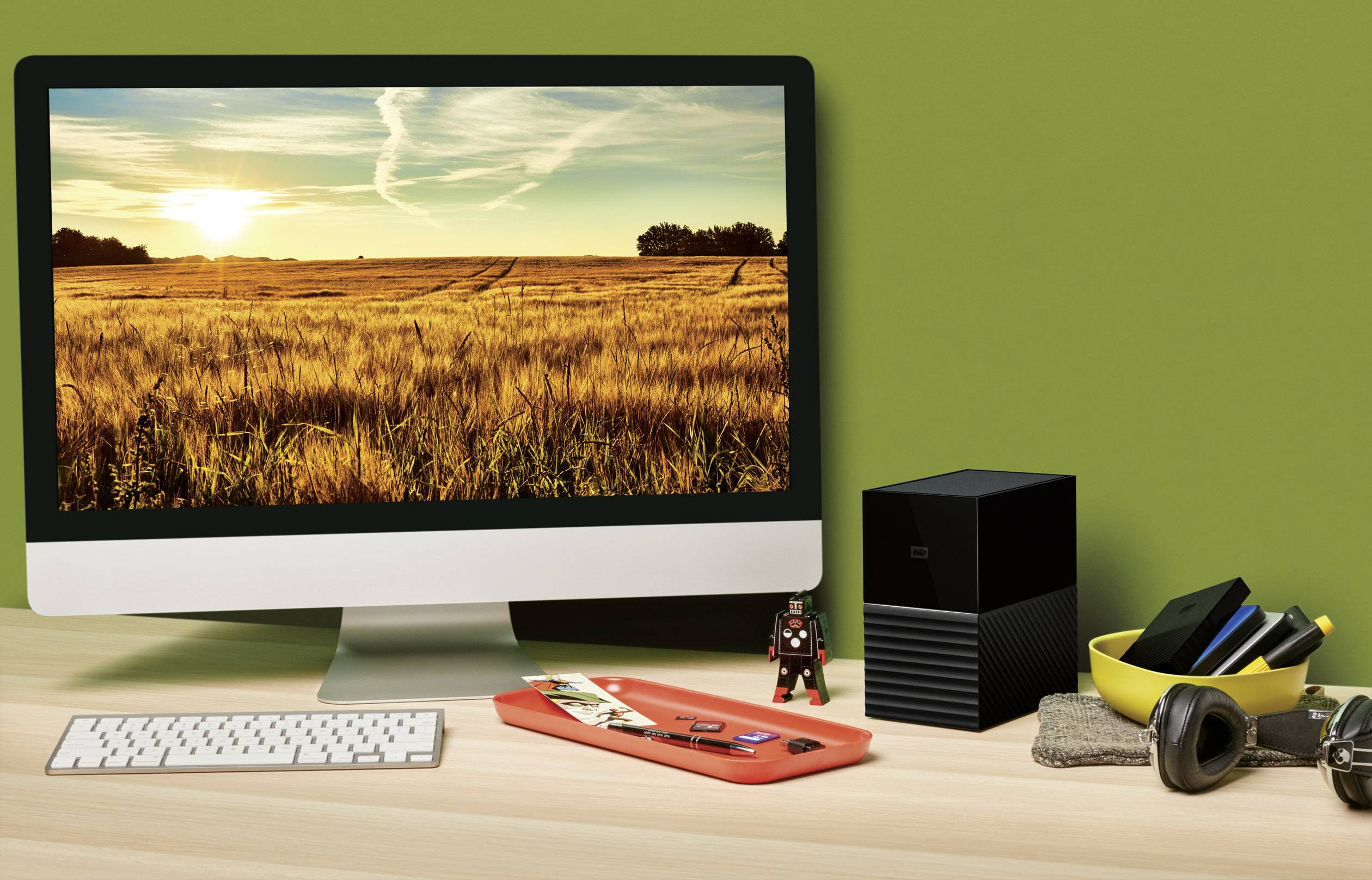 Ein Computerbildschirm auf einem Schreibtisch zeigt eine Landschaft mit einem gelben Weizenfeld im Sonnenuntergang. Daneben liegen Schreibtischutensilien.