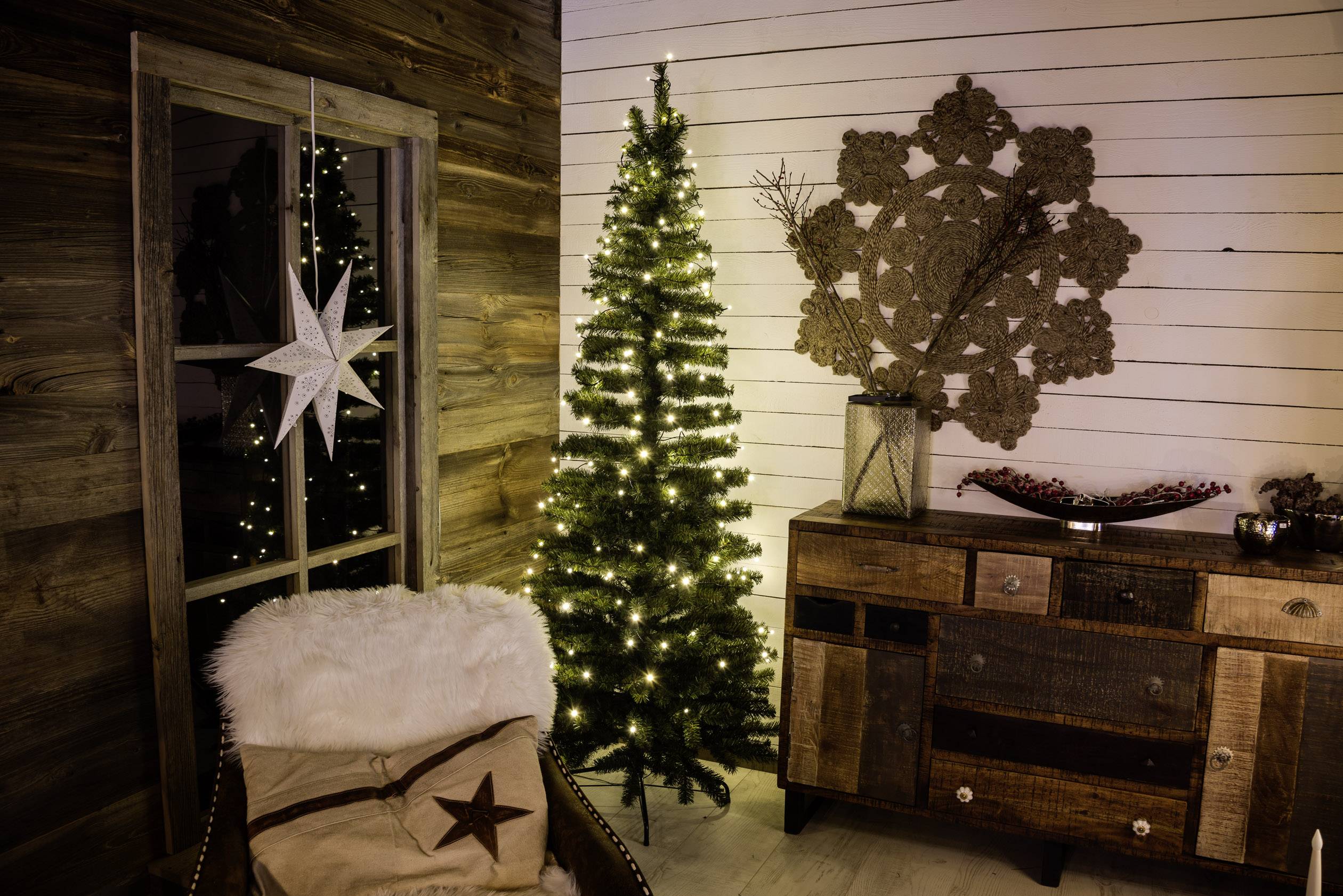 Ein gemütlicher Raum mit einem dekorierten, beleuchteten Weihnachtsbaum neben einem rustikalen Sideboard. Ein Stern hängt am Fenster.