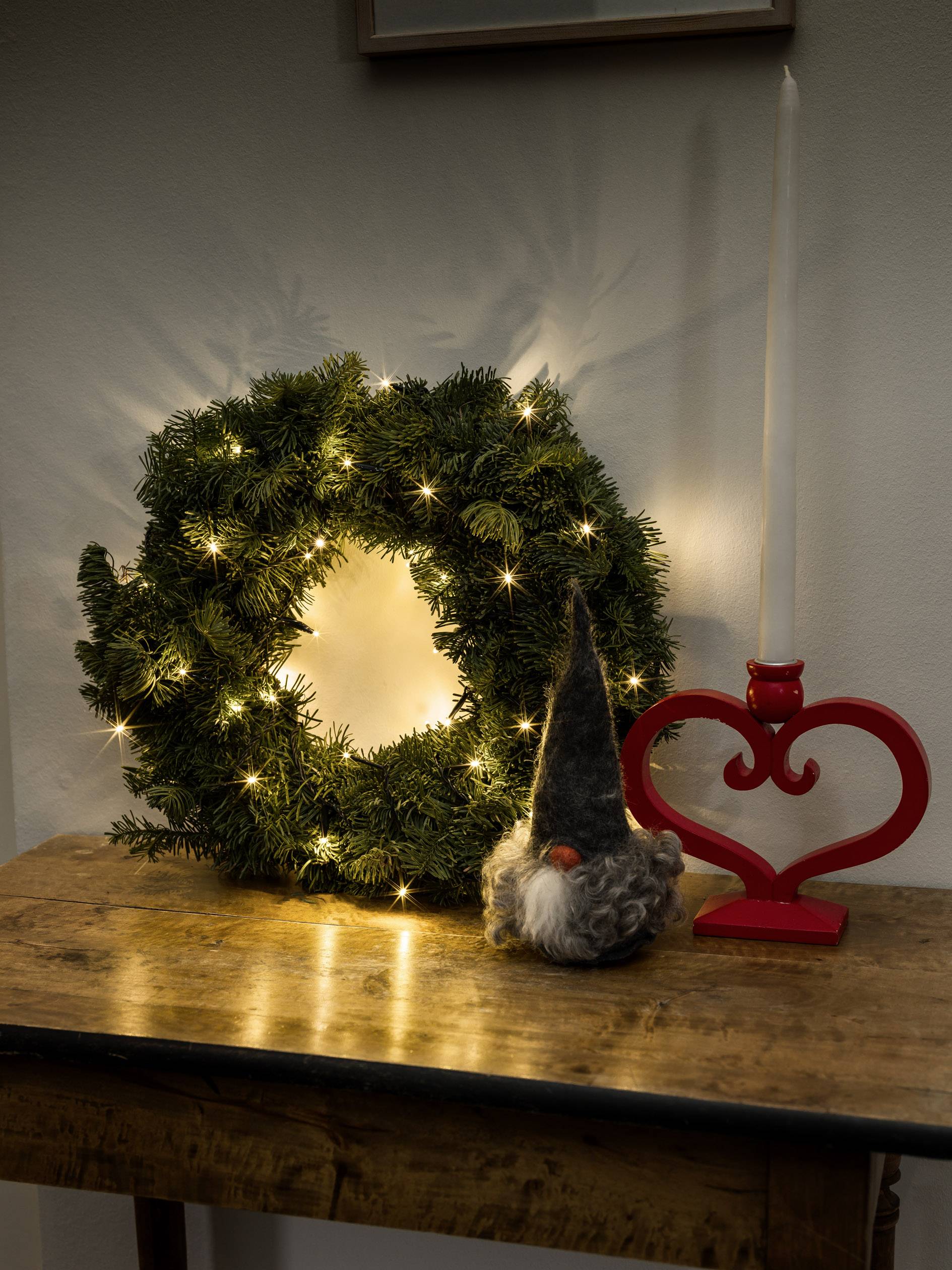 Ein leuchtender Weihnachtskranz aus Tannenzweigen neben einer roten Herzdekoration und einem kleinen Weihnachtswichtel auf einem Tisch.