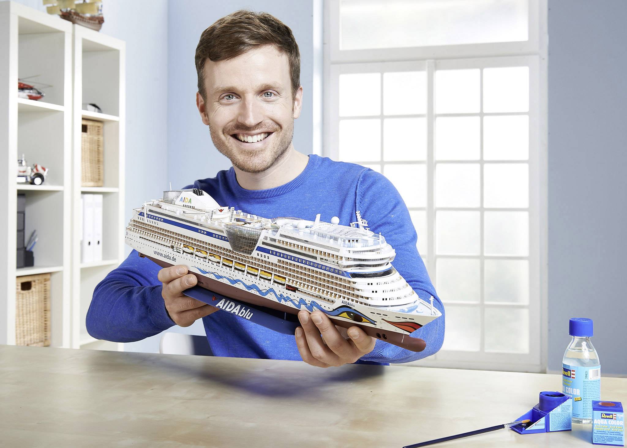 Ein Mann hält ein Modell eines Kreuzfahrtschiffs mit der Aufschrift 'AIDAblu' in einem hellen Raum. Im Hintergrund ein Fenster und Regale.