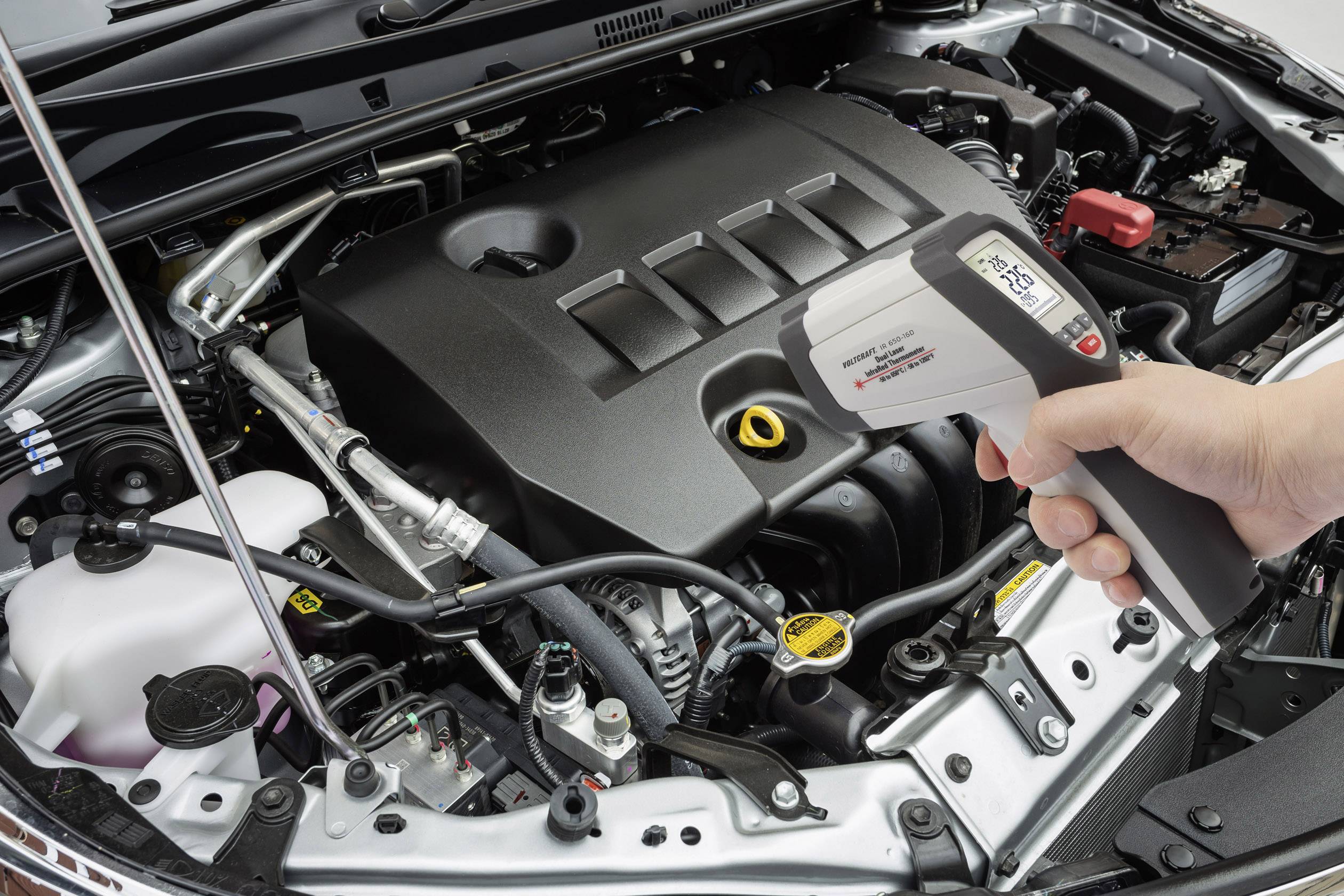 Ein Automechaniker misst mit einem Infrarot-Thermometer die Temperatur eines Motors, der in einem offenen Motorraum eines Autos zu sehen ist.