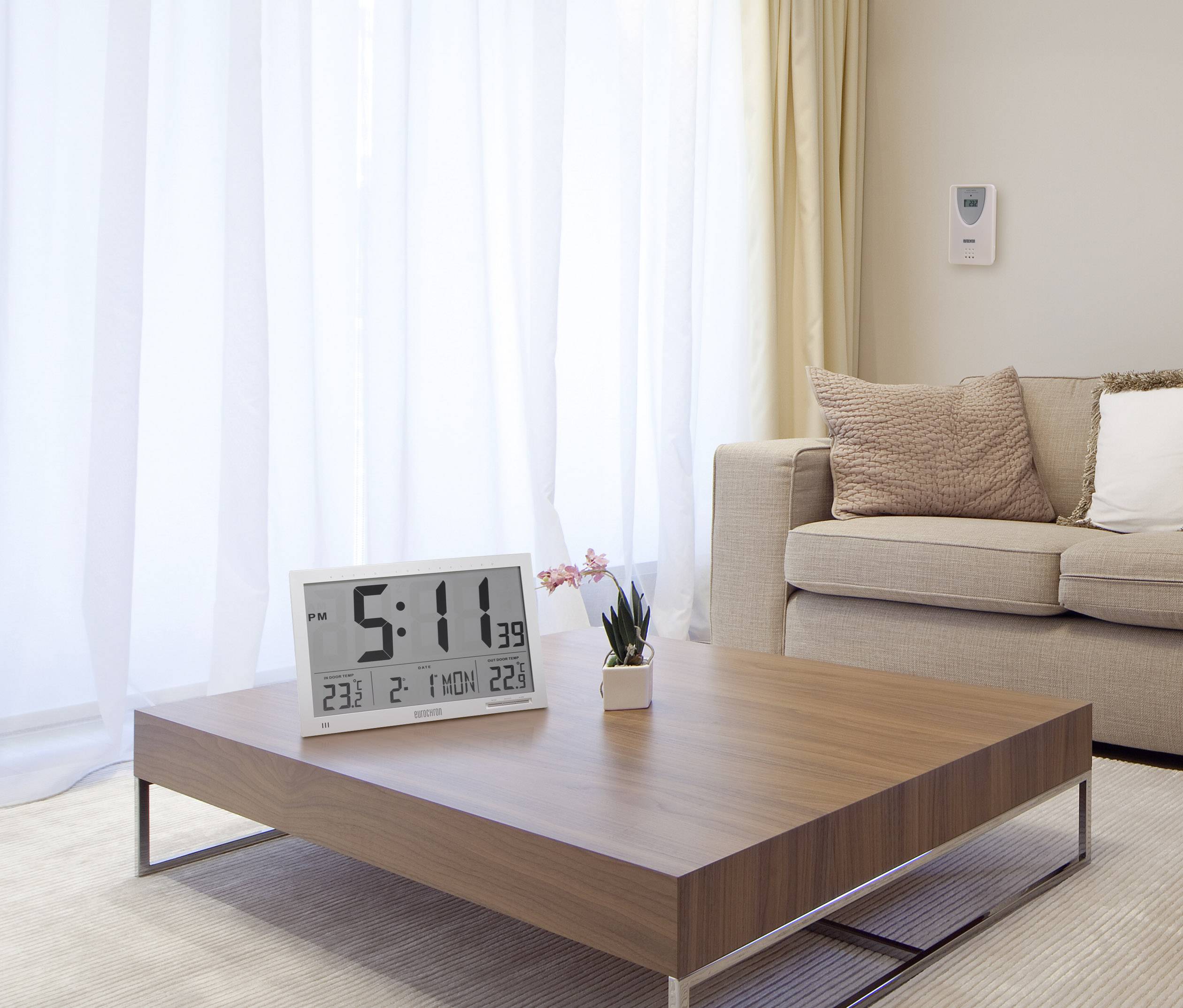 Ein modernes Wohnzimmer mit einem Holztisch, auf dem ein großer digitaler Wecker steht. Ein Sofa mit Kissen ist im Hintergrund.