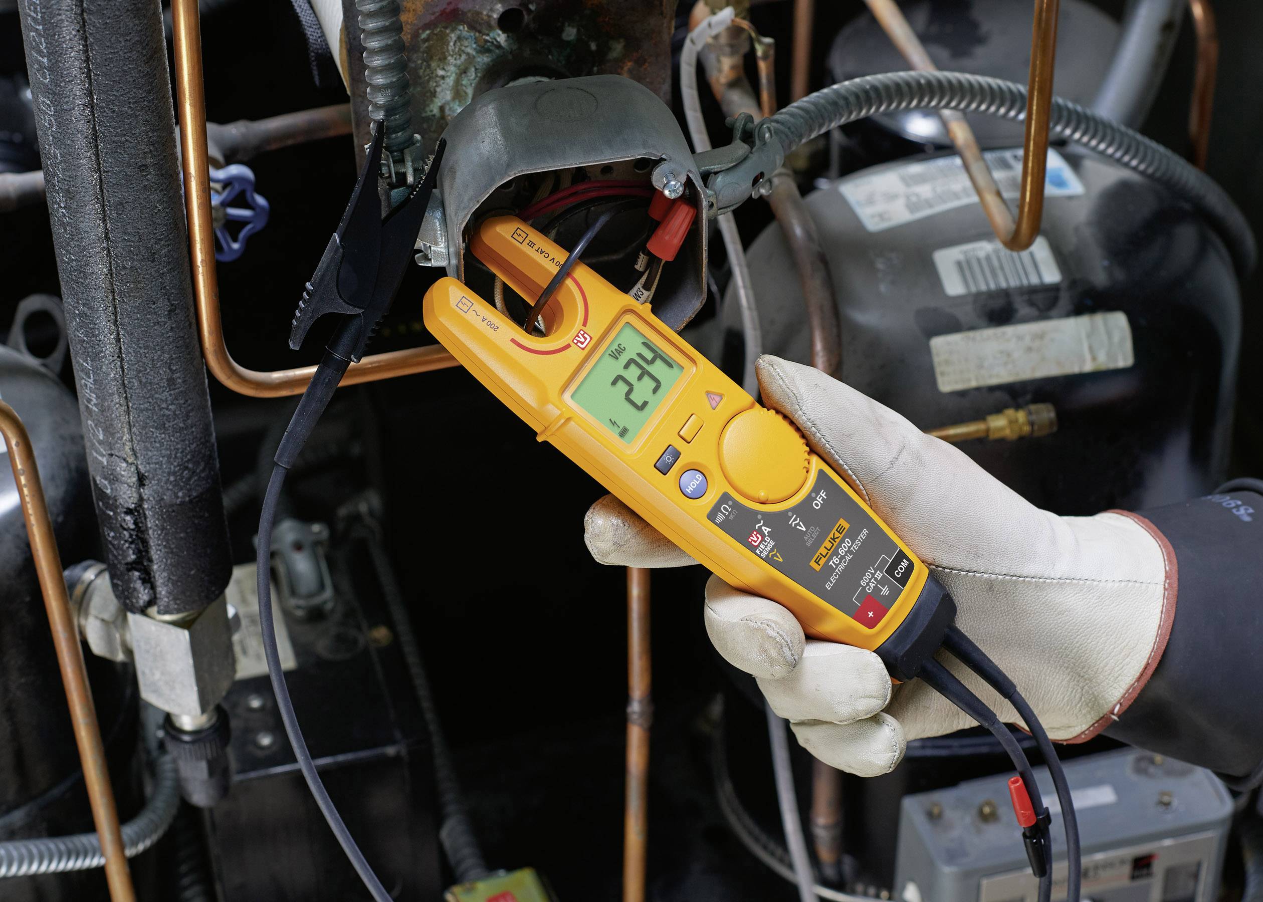 Eine Person hält ein digitales Multimeter, das 234 Volt an einer elektrischen Anlage anzeigt. Hintergrund zeigt Kupferrohre und Kabel.