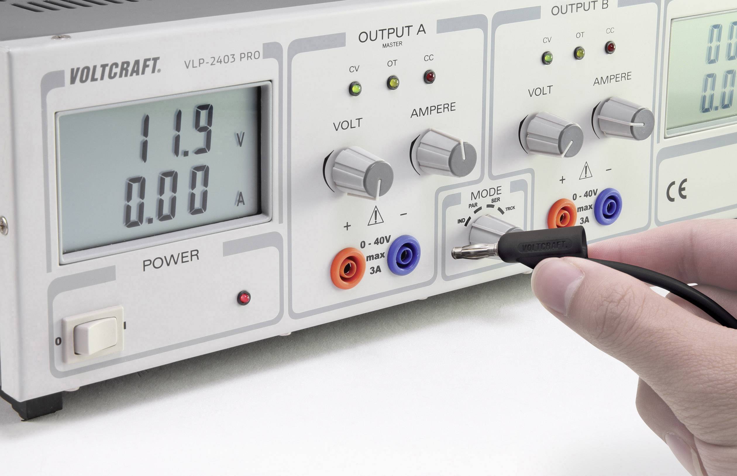 Eine Person bedient ein Spannungsmessgerät mit Anzeige von 11,9 V und 0,00 A. Geräteknöpfe sind blau, orange und graurot.