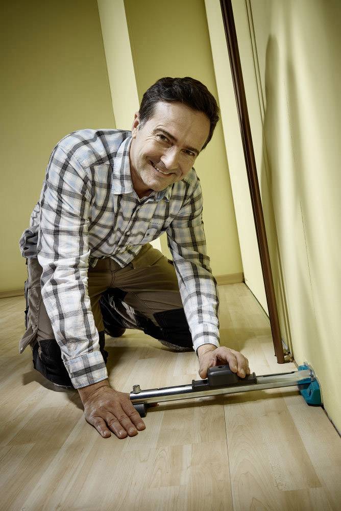 Ein Mann kniet auf einem Holzboden und verwendet ein Werkzeug, um die Wand abzumessen.