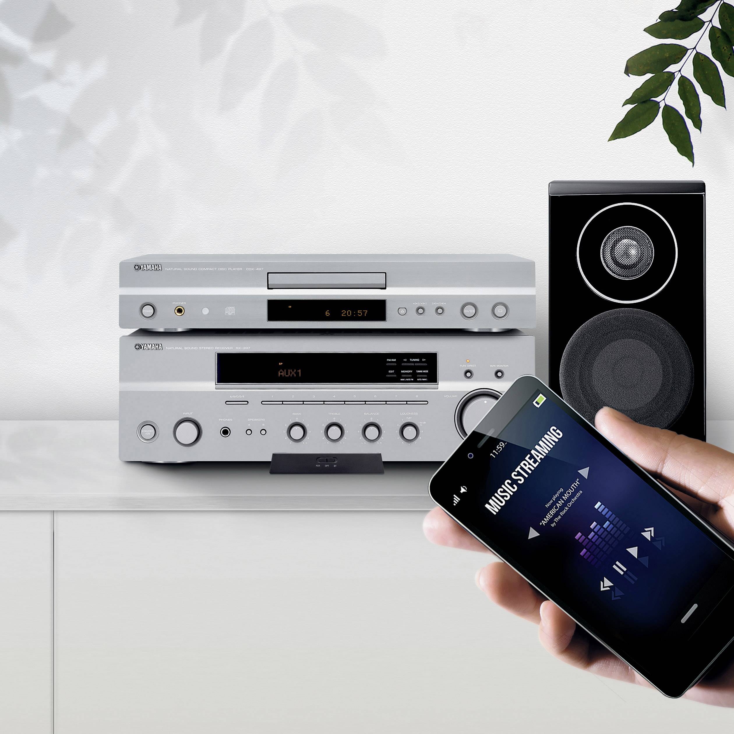 Eine Hand hält ein Smartphone, das Musik-Streaming zeigt, im Vordergrund. Im Hintergrund steht ein Stereoanlagen-Setup mit Lautsprecher.