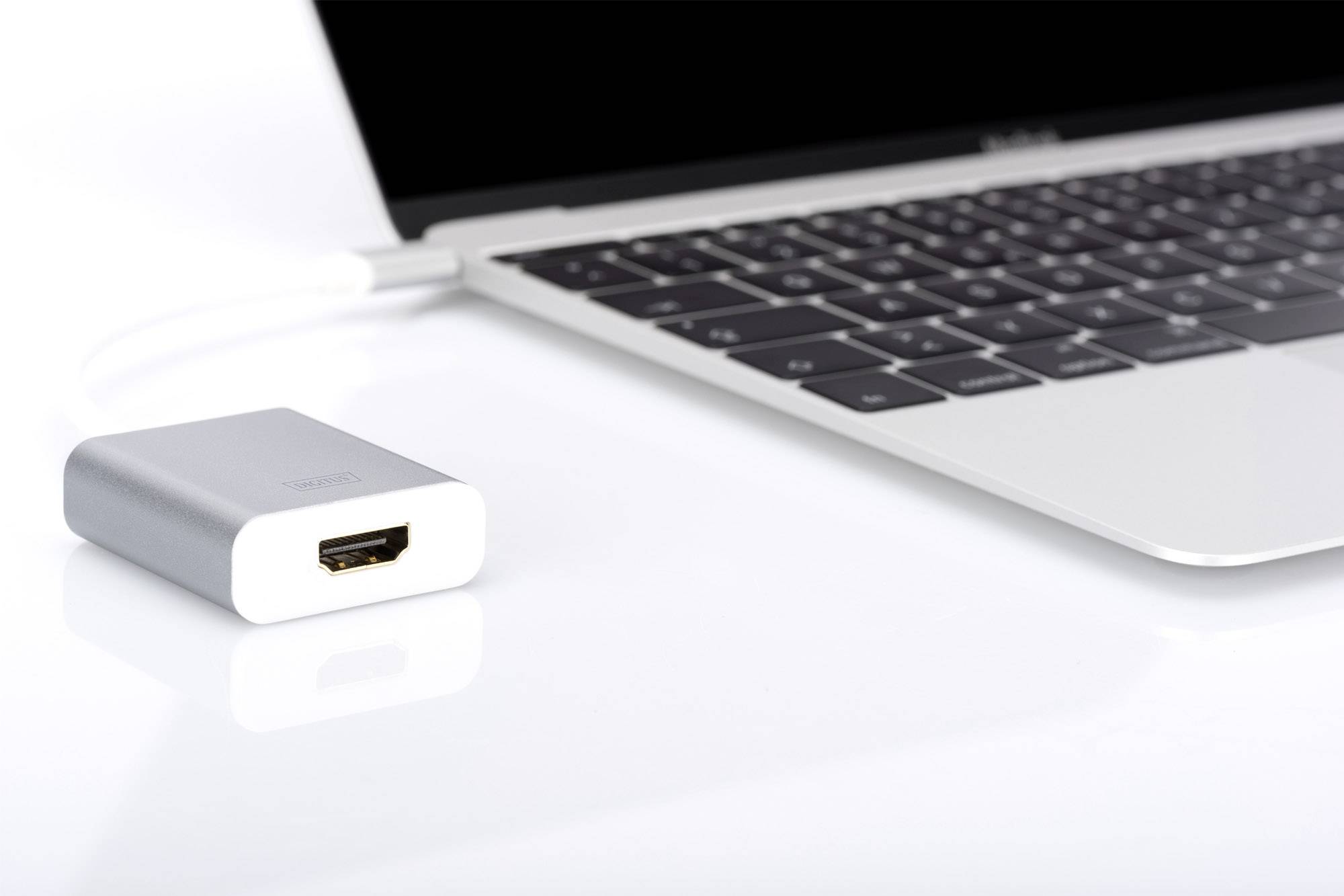 Ein silberner HDMI-Adapter liegt neben einem Laptop mit schwarzer Tastatur auf einem weißen Tisch. Adapter ist nicht angeschlossen.