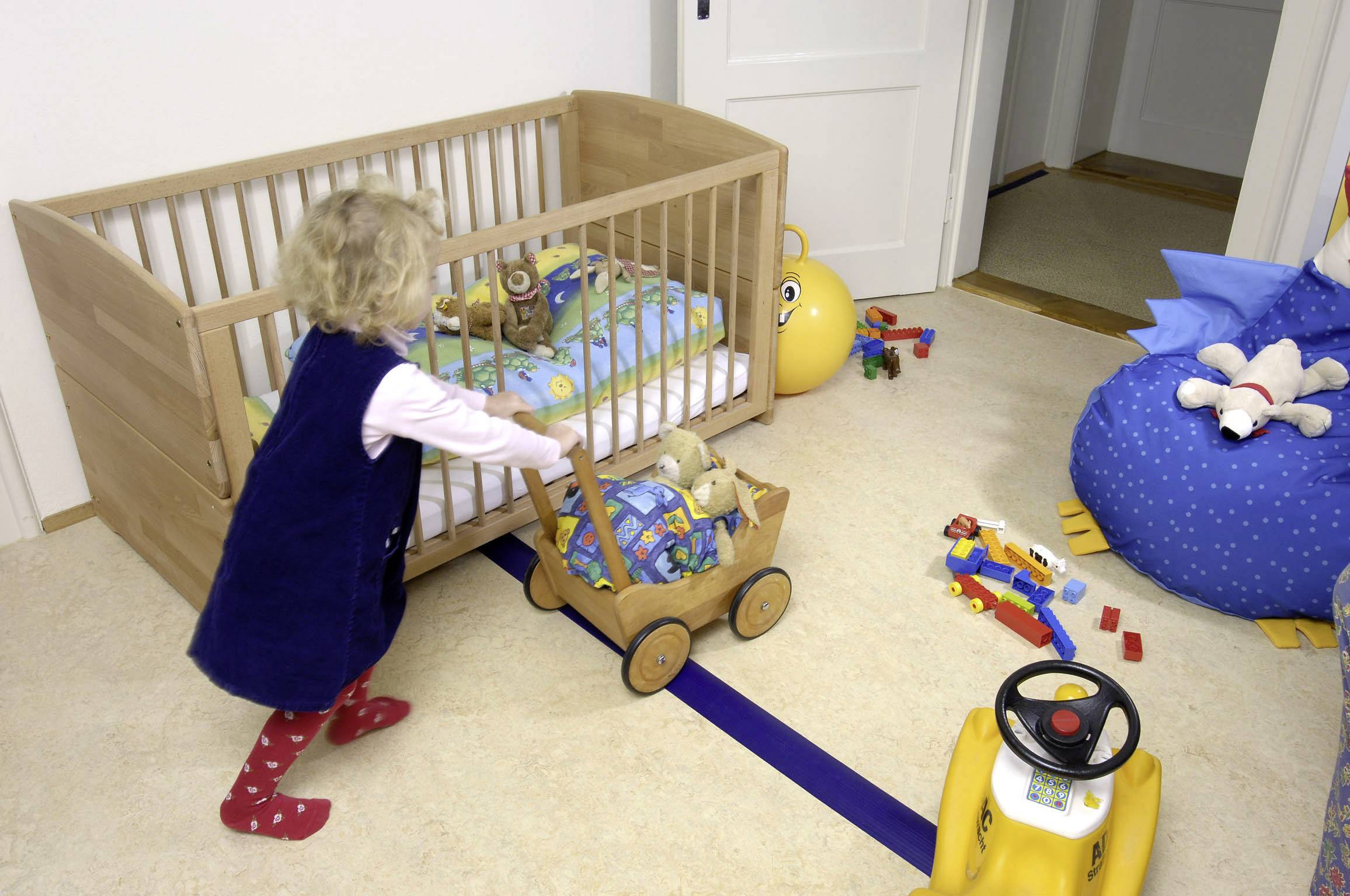 Ein kleines Kind spielt in einem Kinderzimmer. Es schiebt einen Holzwagen mit Spielzeug. Im Hintergrund steht ein Gitterbett.