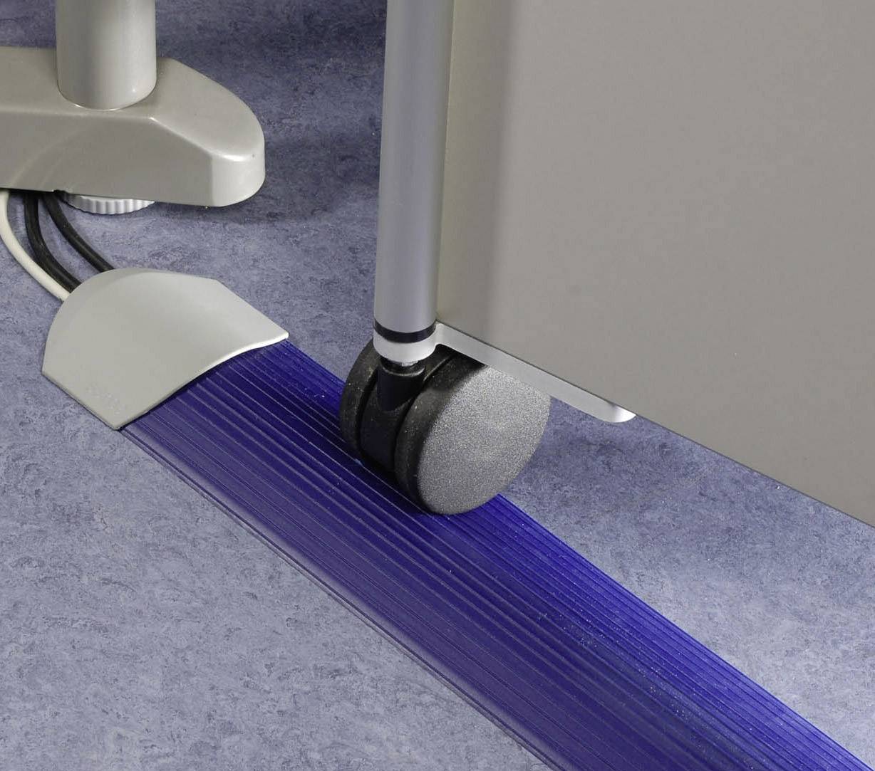 Bürostuhlrolle auf einem blauen Kabelkanal, der Kabel am Boden ordnet und schützt. Hintergrund: grauer Bodenbelag und Möbel.