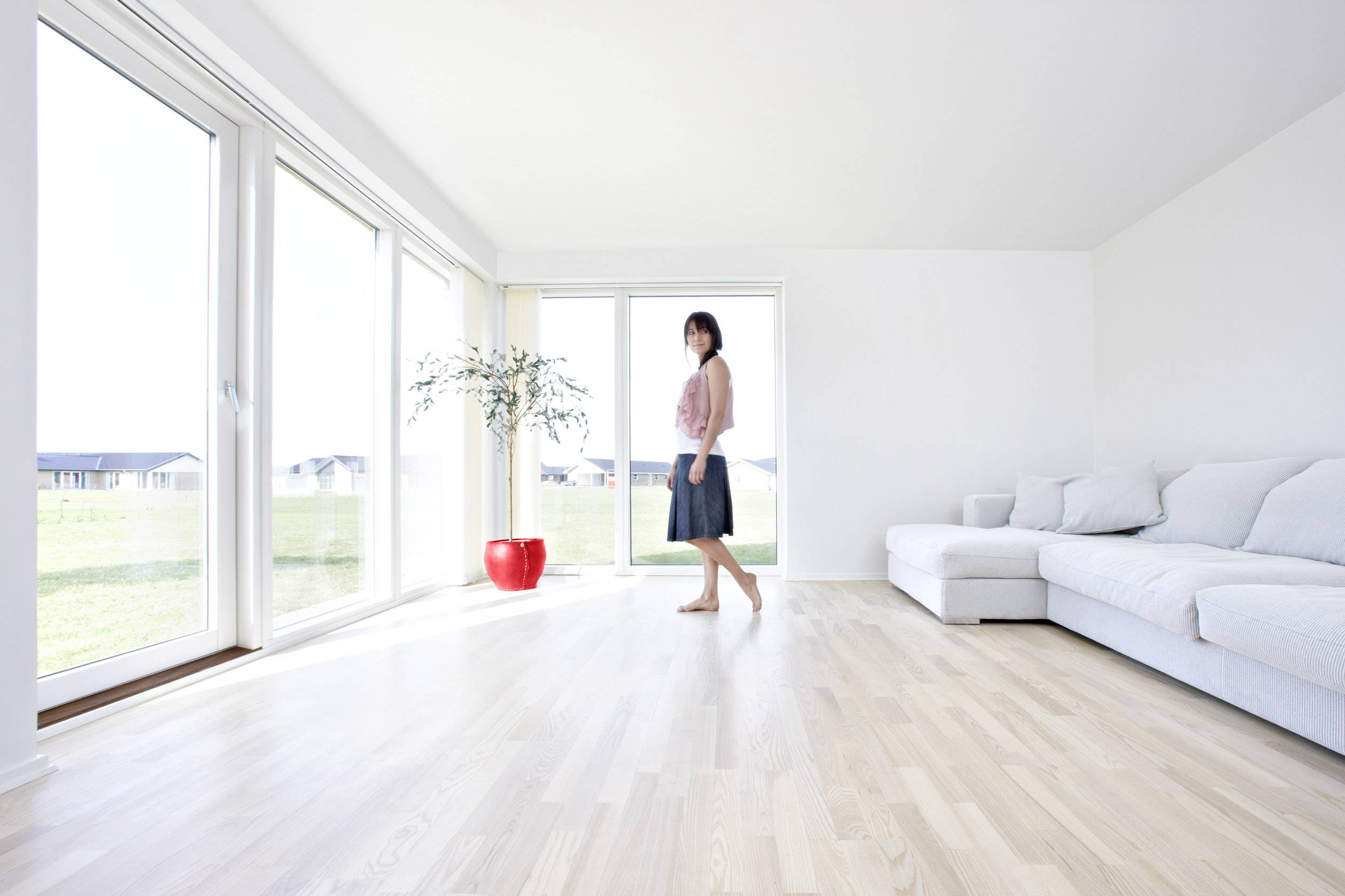 Eine Frau steht in einem großen, hellen Wohnzimmer mit Holzboden. Große Fenster lassen viel Licht herein. Links steht eine Pflanze.