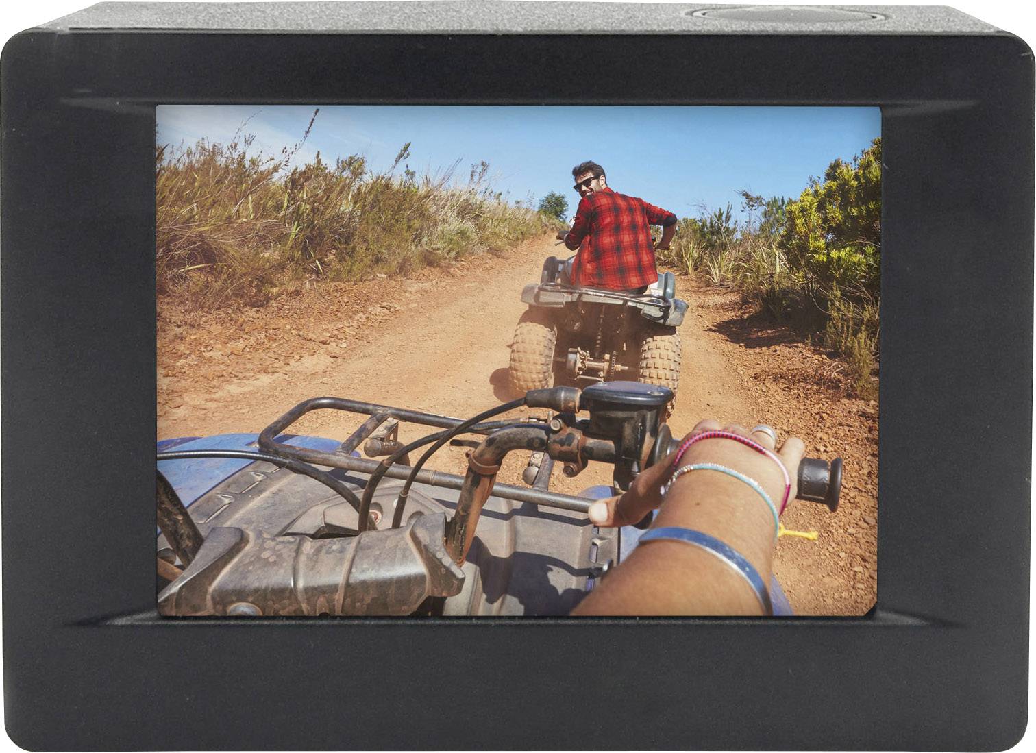 Eine Person in rotem Hemd fährt auf einem Quad über einen staubigen Weg in einer ländlichen Gegend, gefolgt von einer weiteren Person.