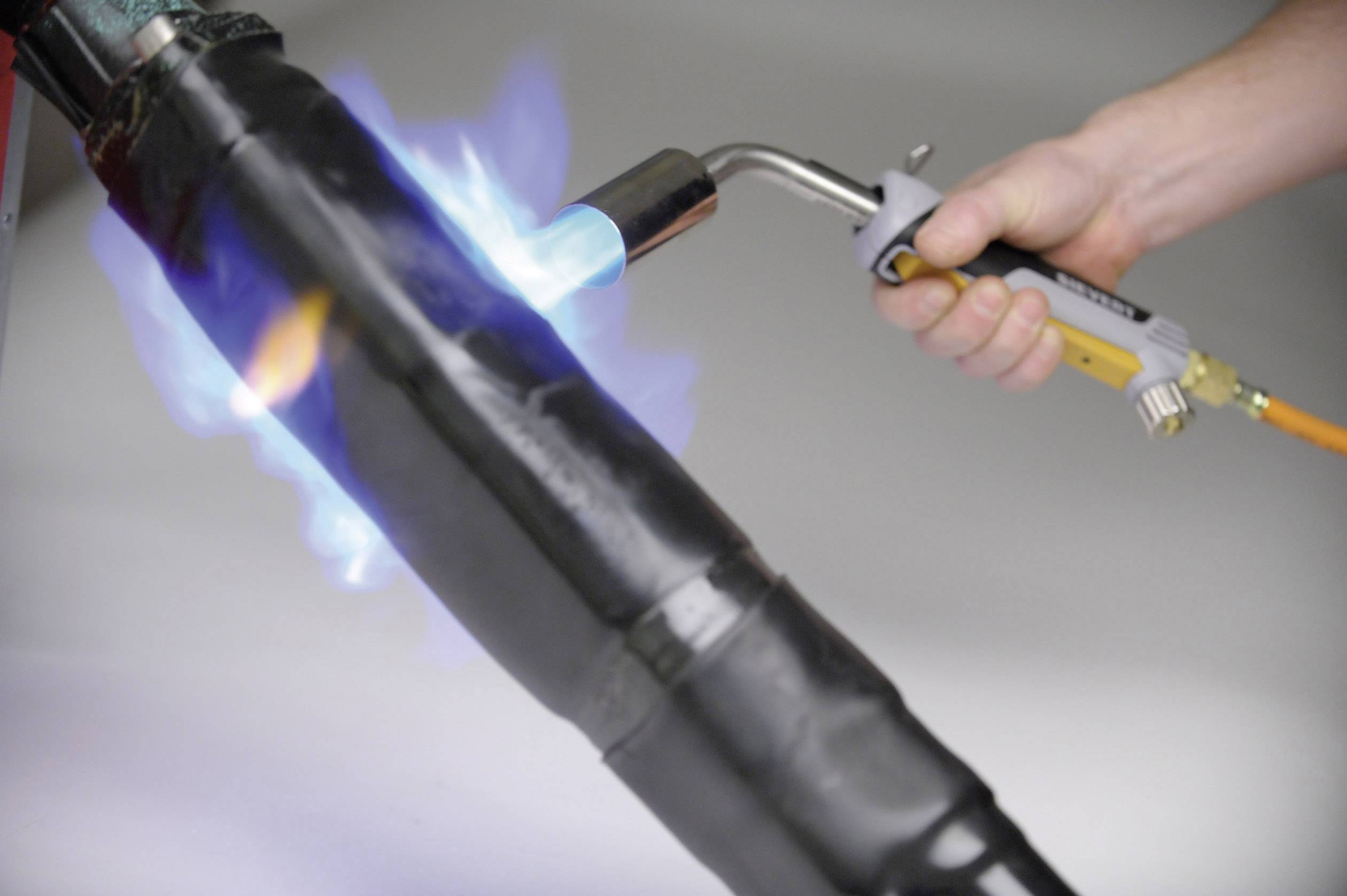 Eine Hand hält einen Gasbrenner, der eine blaue Flamme erzeugt, um eine schwarze Rohroberfläche zu erhitzen.