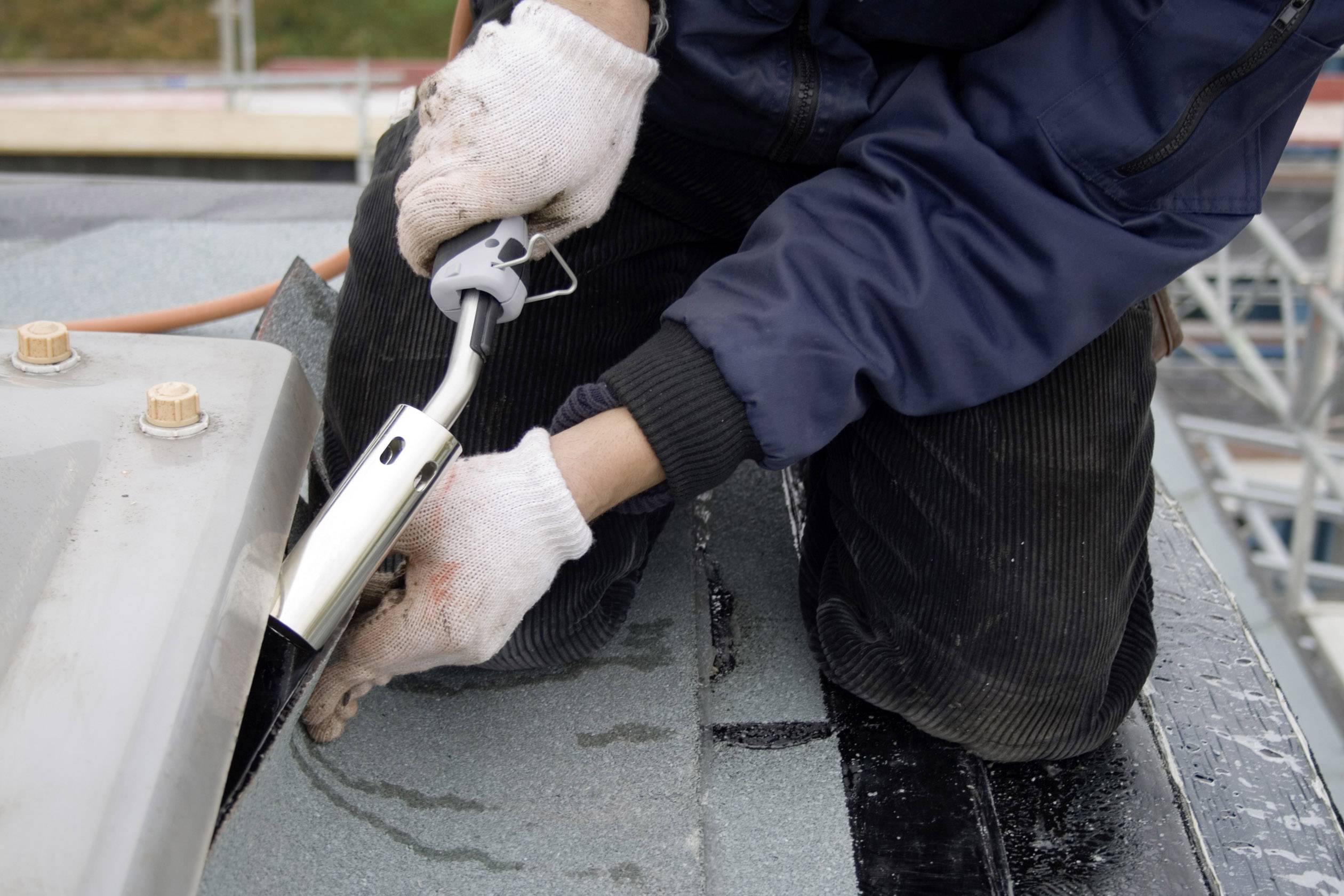 Eine Person mit weißen Handschuhen benutzt ein Werkzeug, um Dachabdichtungsmaterial zu bearbeiten. Die Umgebung ist eine Baustelle.