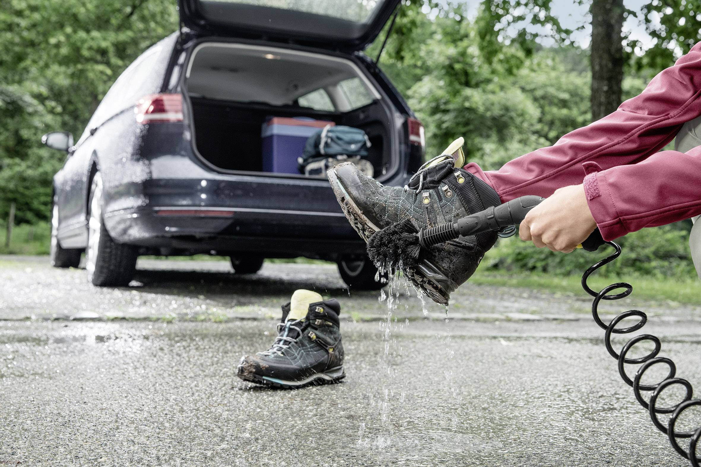 Eine Person reinigt schmutzige Wanderschuhe mit einem Wasserschlauch neben einem Auto. Der Kofferraum ist geöffnet, zeigt Gepäck.