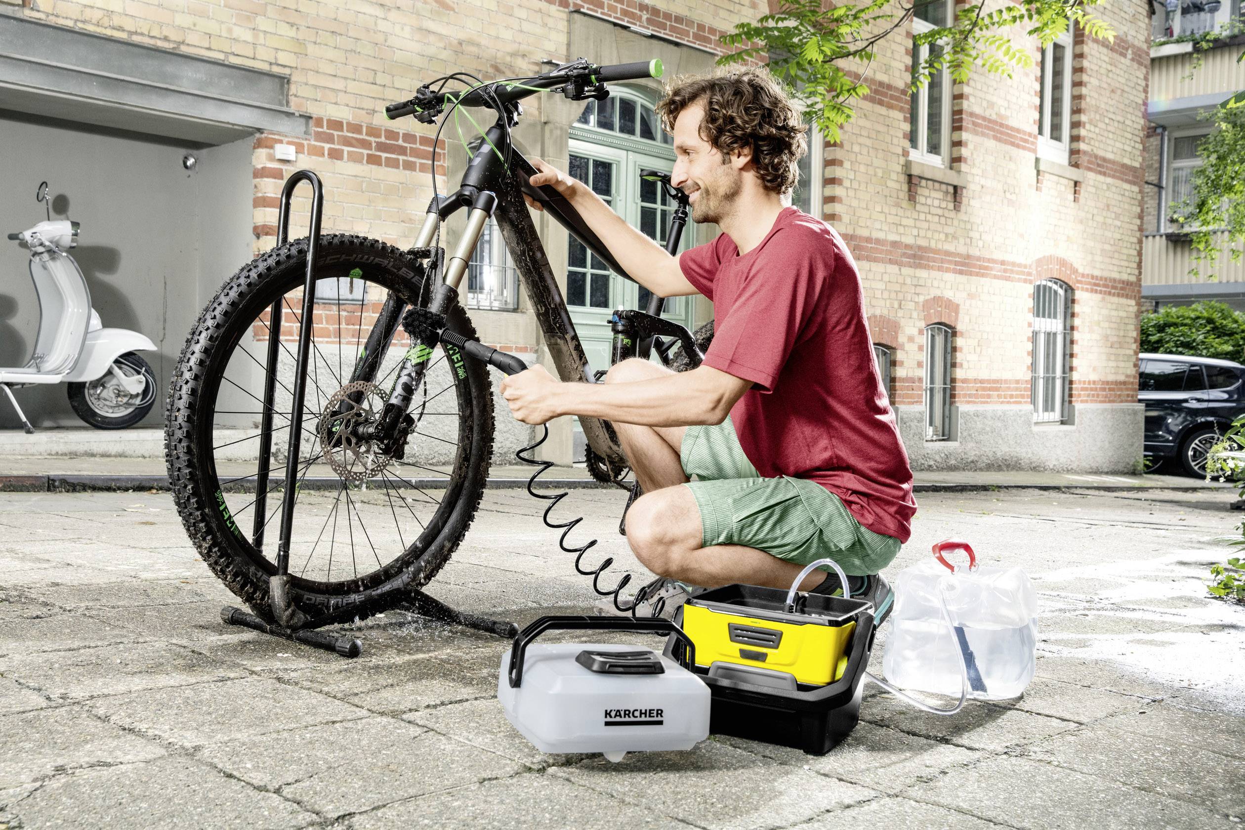 Ein Mann reinigt ein Mountainbike mit einem tragbaren Hochdruckreiniger auf einem gepflasterten Hof vor einem Backsteingebäude.