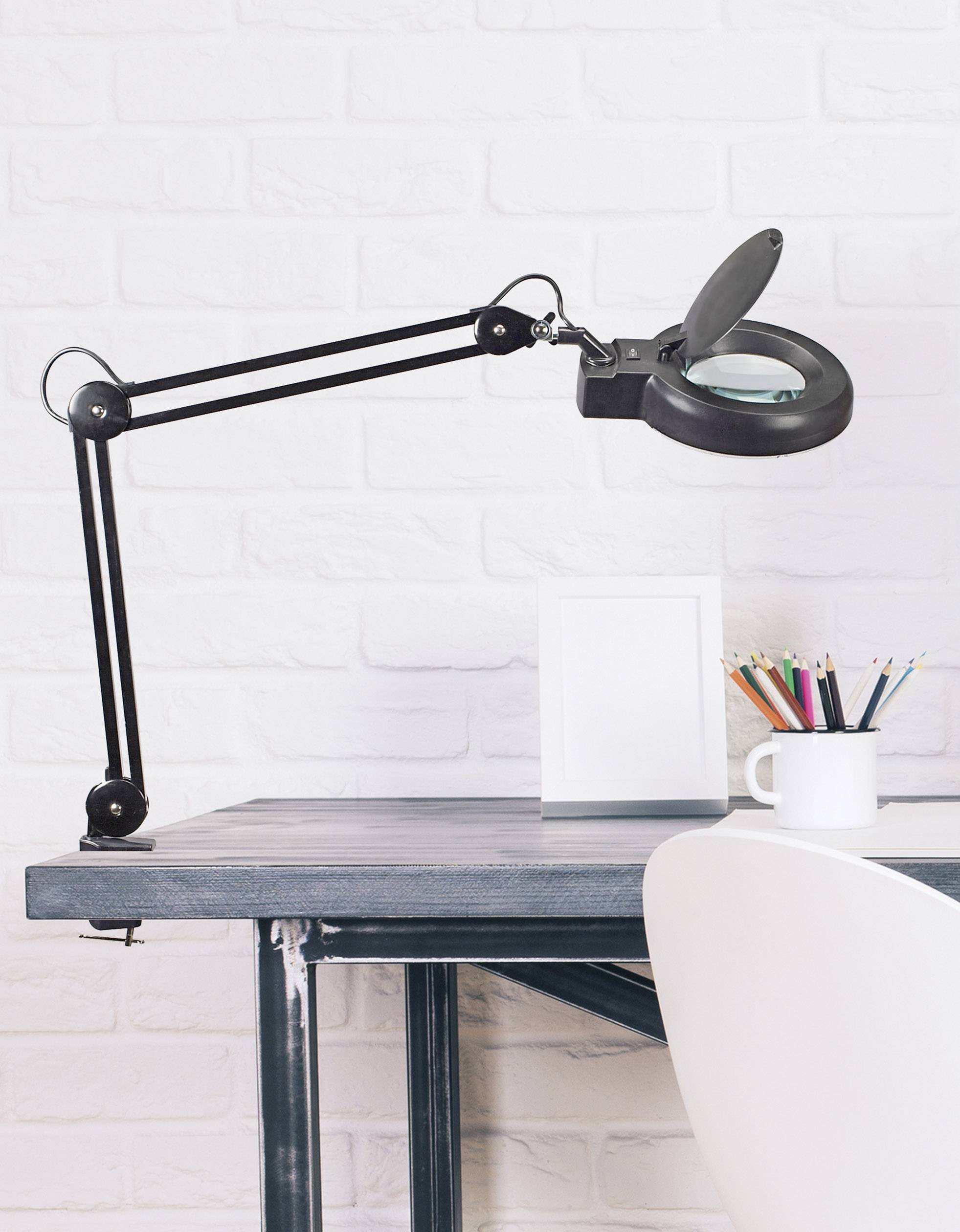 Schreibtisch mit einer schwarzen Tischlampe mit Lupe, leeres Bilderrahmen, bunte Stifte in einer Tasse, vor einer weißen Ziegelwand.