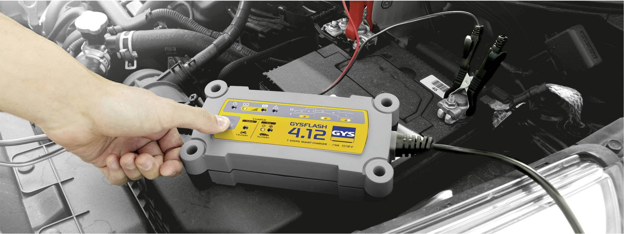 Eine Person schließt ein Ladegerät an eine Autobatterie an. Das Ladegerät zeigt '4.12' an und ist mit Klemmen verbunden.