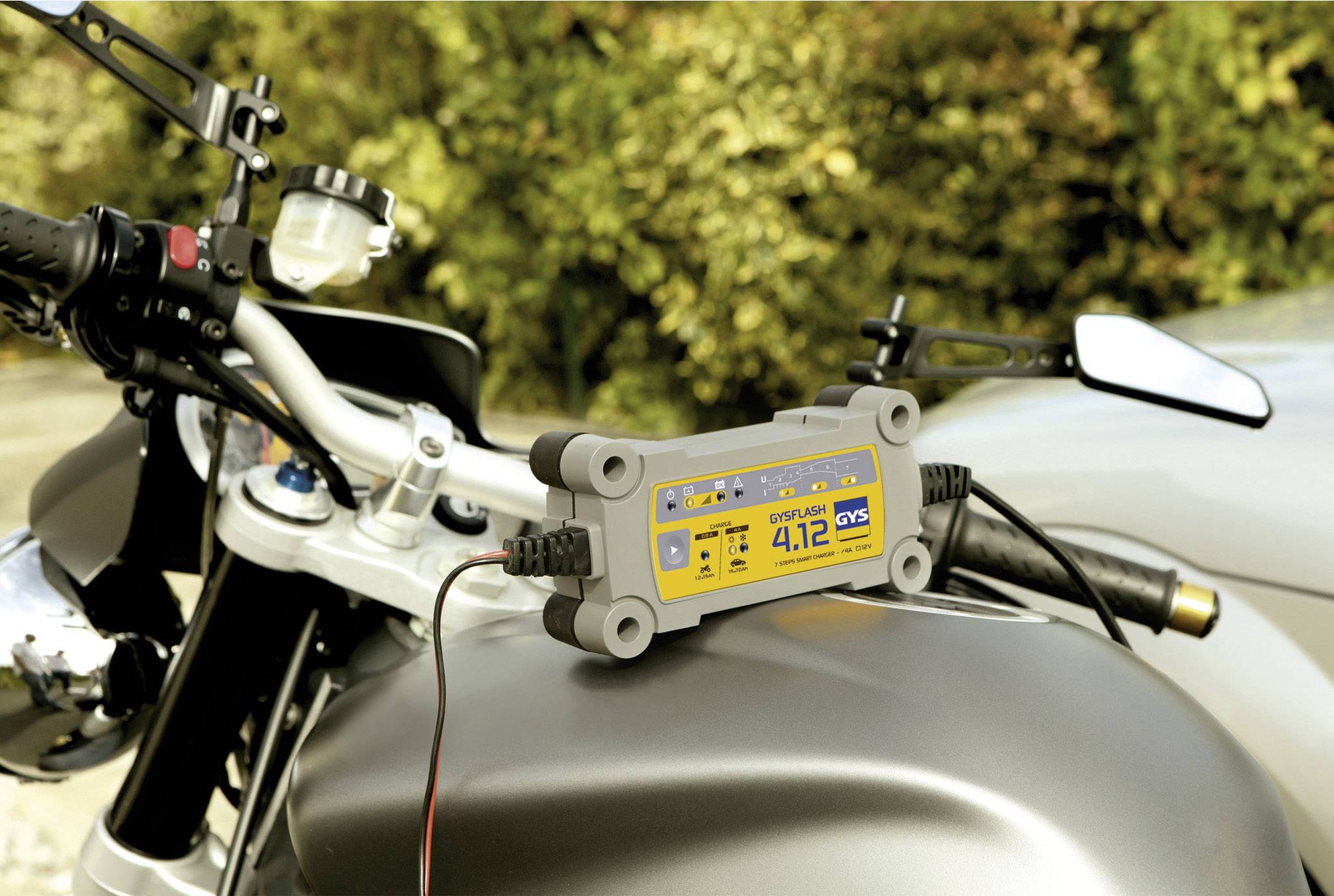 Ein Batterieladegerät ist an ein Motorrad angeschlossen, das auf einer Straße mit Bäumen im Hintergrund steht.