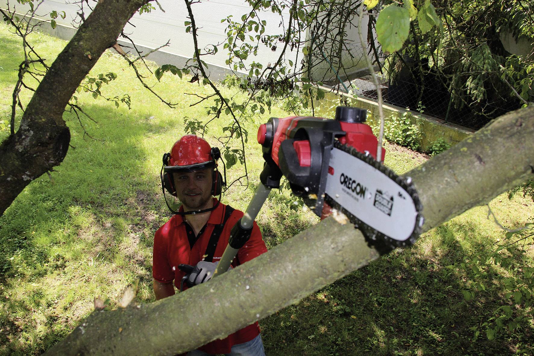 Ein Mann mit rotem Schutzhelm und Brille schneidet mit einer elektrischen Astsäge einen dicken Ast von einem Baum ab.