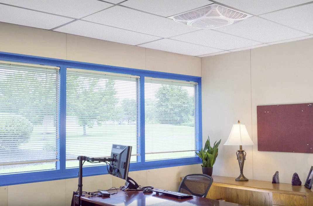 Büro mit Schreibtisch, Stuhl, Computerbildschirm, Lampe und Pflanze. Große Fenster bieten Blick ins Freie mit Bäumen und Rasenfläche.