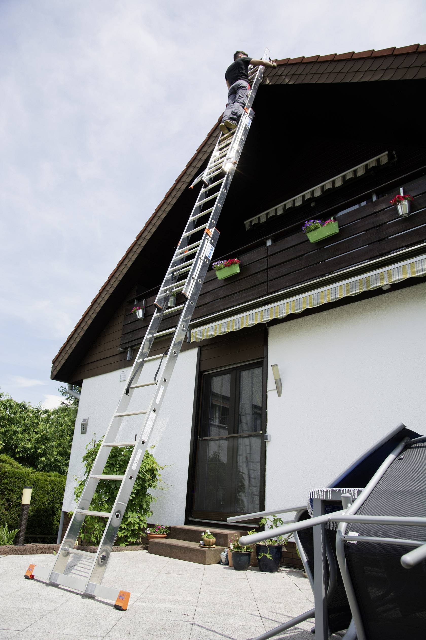 Eine Person steht auf einer hohen Leiter und arbeitet am Dach eines Hauses. Die Umgebung zeigt eine Terrasse mit Pflanzen.