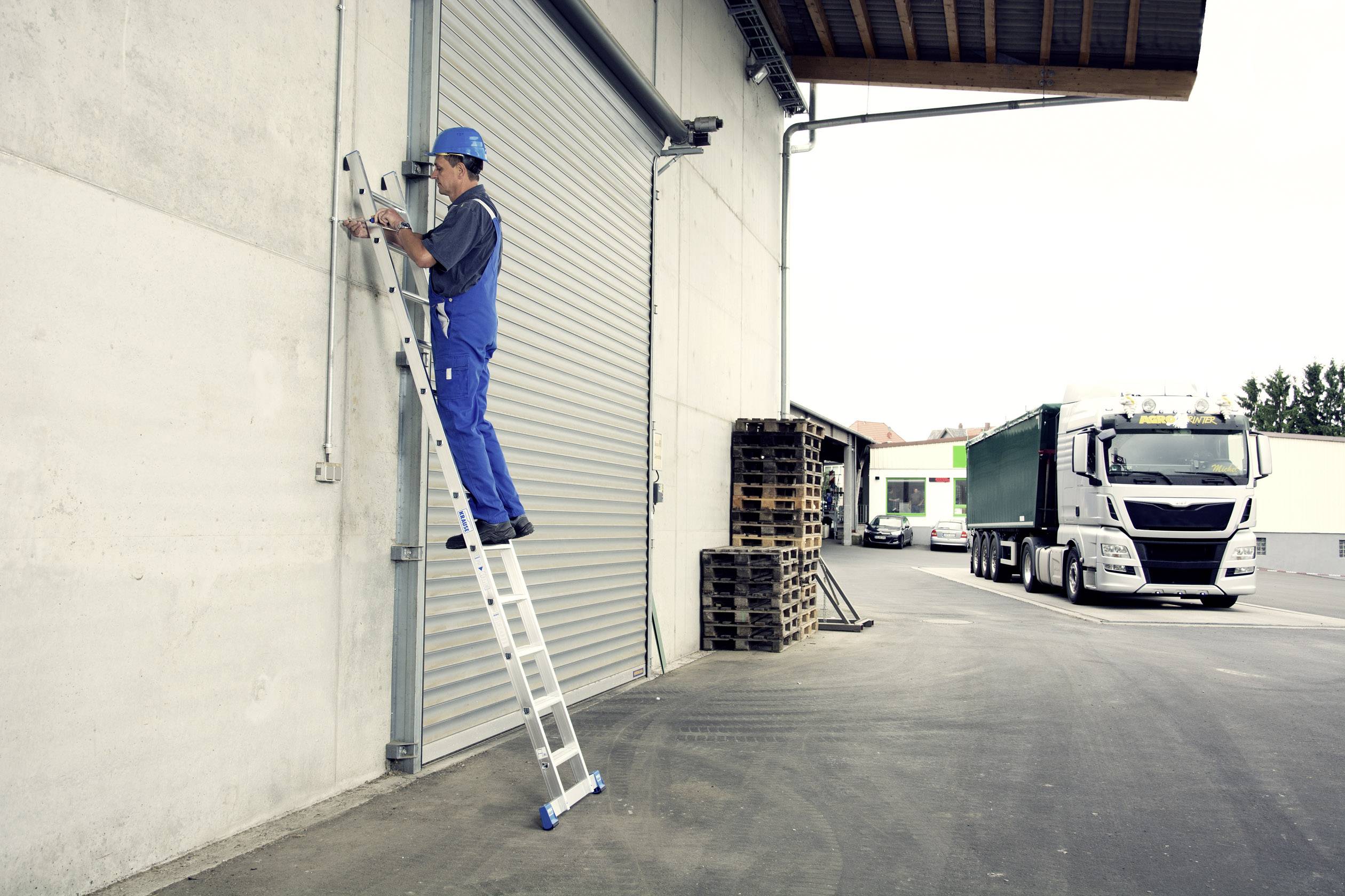 Ein Arbeiter auf einer Leiter repariert ein Industrietor, während ein Lastwagen im Hintergrund parkt. Paletten sind an der Wand gestapelt.