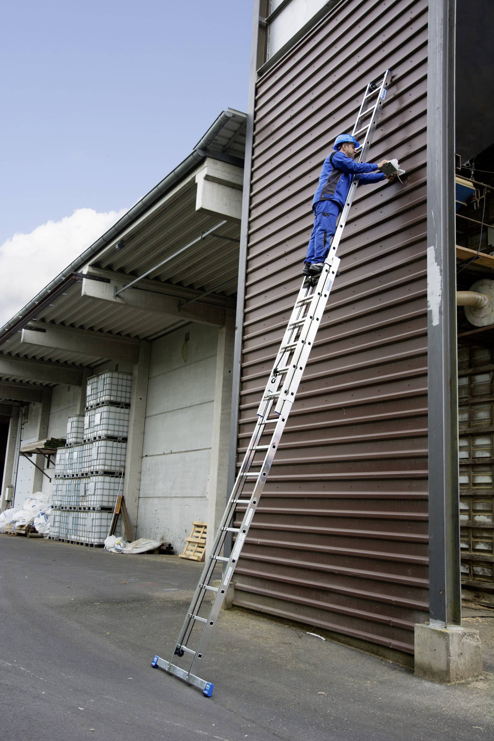 Ein Arbeiter in blauer Uniform steht oben auf einer hohen Leiter, befestigt mit Werkzeug Metallpaneele an einer Gebäudewand.