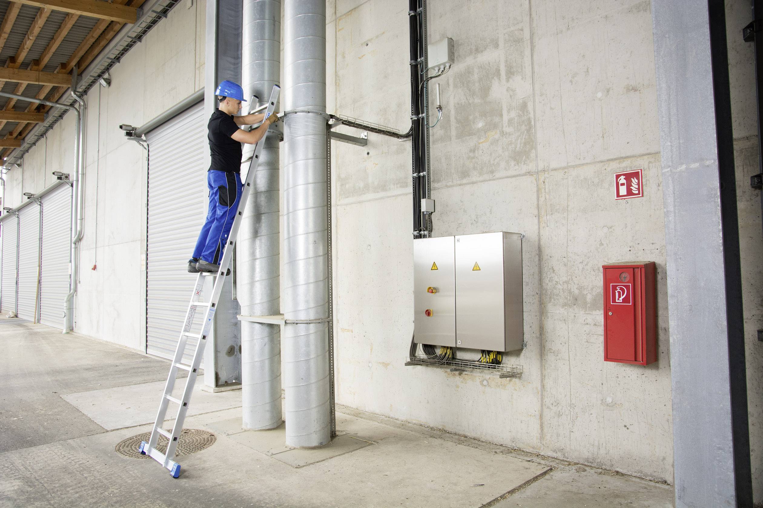 Ein Arbeiter steht auf einer Leiter und arbeitet an Rohren an einer Gebäudewand. Sicherheitsausrüstung, wie Helm, wird getragen.