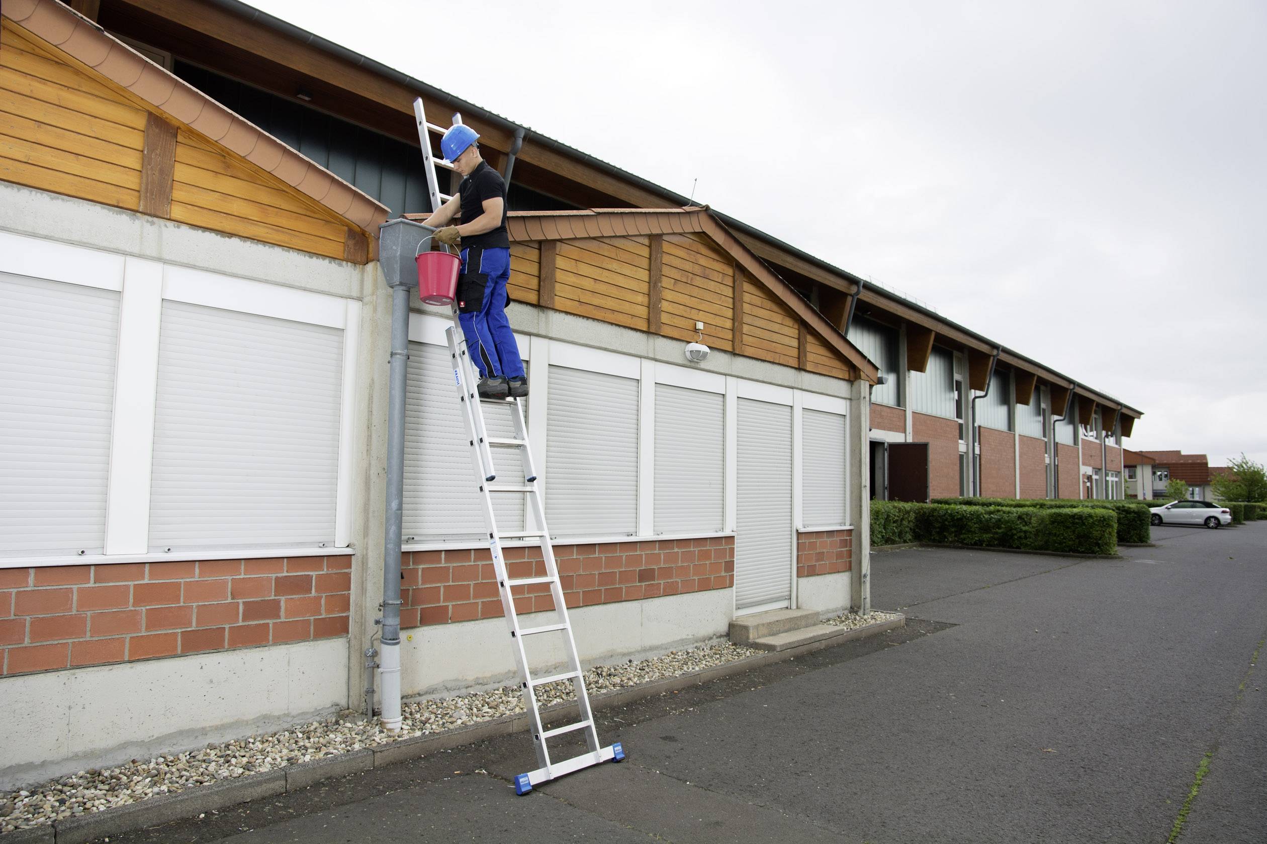 Eine Person auf einer Leiter reinigt eine Dachrinne an einem Gebäude. Sie trägt Sicherheitsausrüstung und stützt sich auf den Rinnenrand.