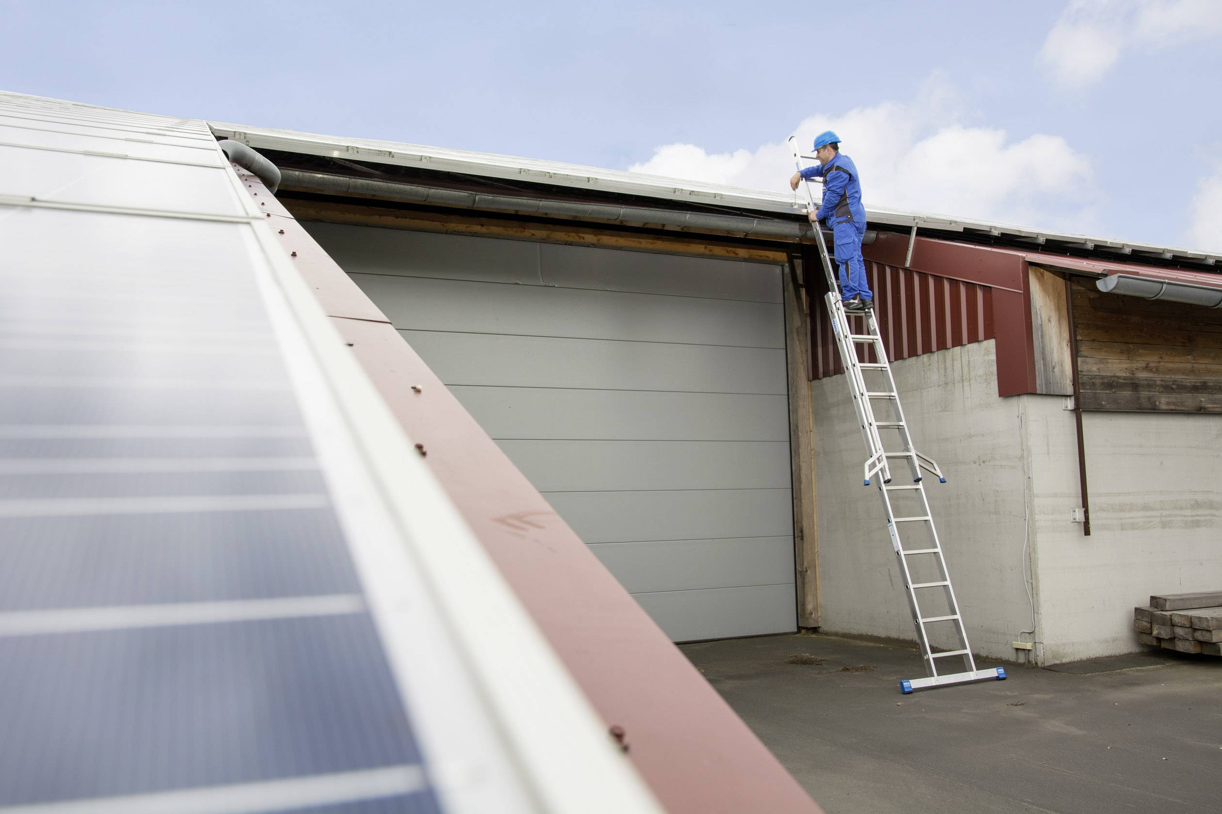 Ein Arbeiter in blauer Uniform auf einer Leiter repariert das Dach eines Lagerhauses. Links sind Solarpaneele zu sehen.