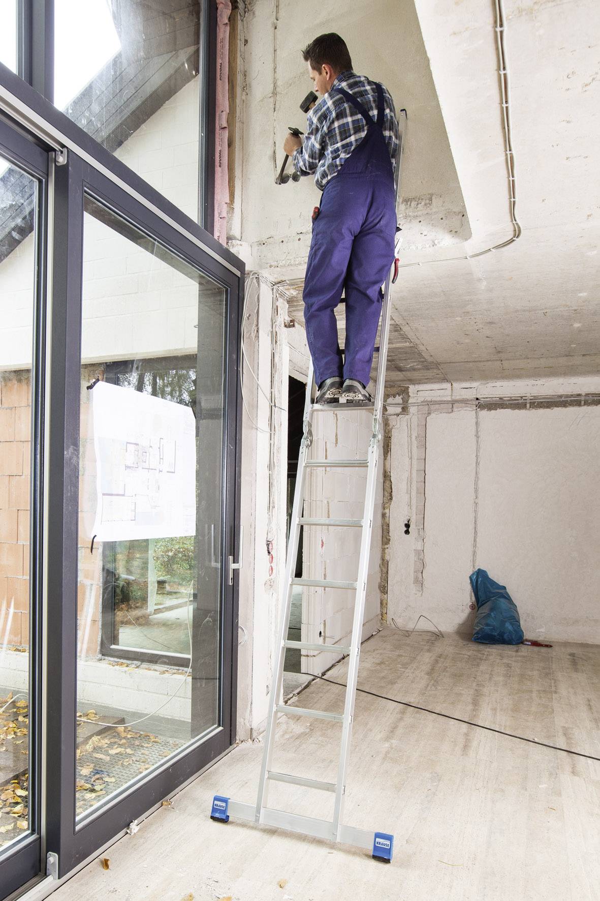 Ein Mann auf einer Leiter arbeitet mit einem Werkzeug an der Decke in einem unfertigen Raum mit großen Fenstern.