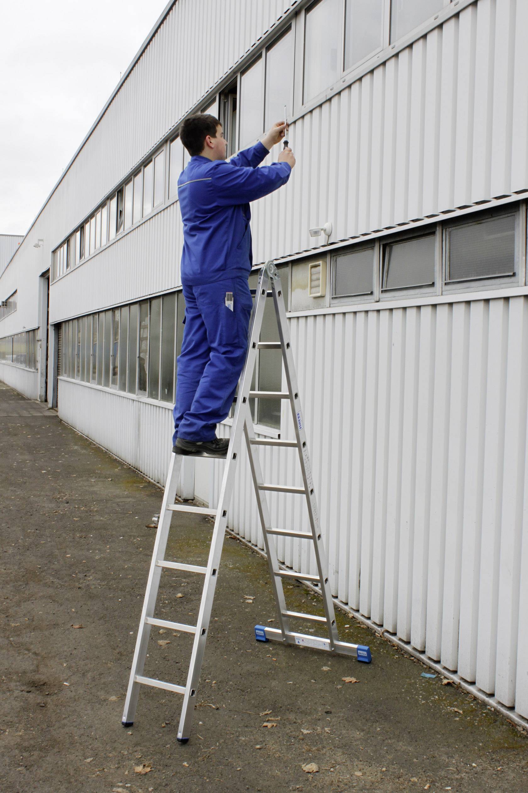 Ein Mann in blauer Arbeitskleidung steht auf einer Leiter und arbeitet an der Außenwand eines Industriegebäudes.