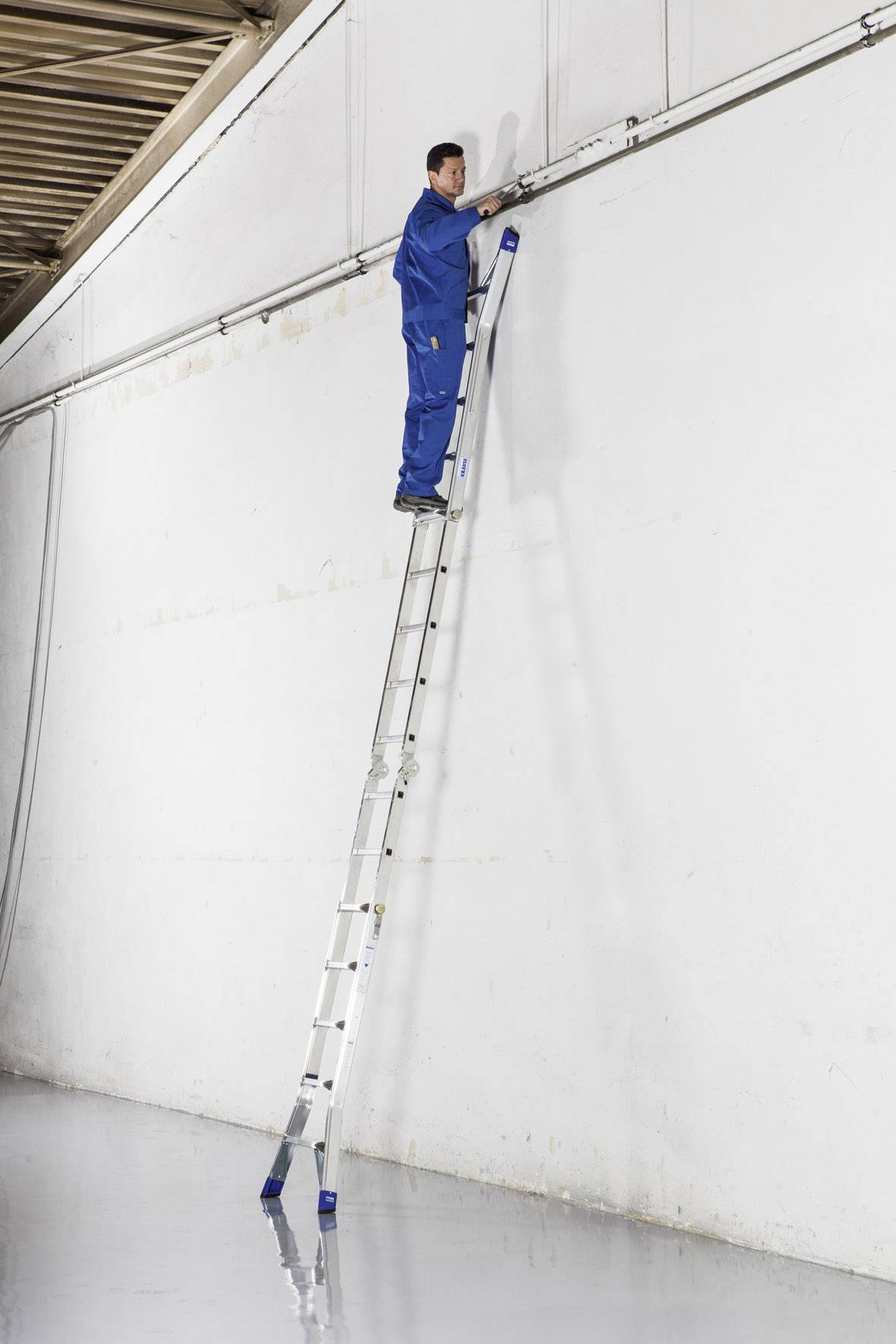 Ein Mann steht oben auf einer langen Leiter, die an einer hohen weißen Wand lehnt, in einem großen Raum mit reflektierendem Boden.