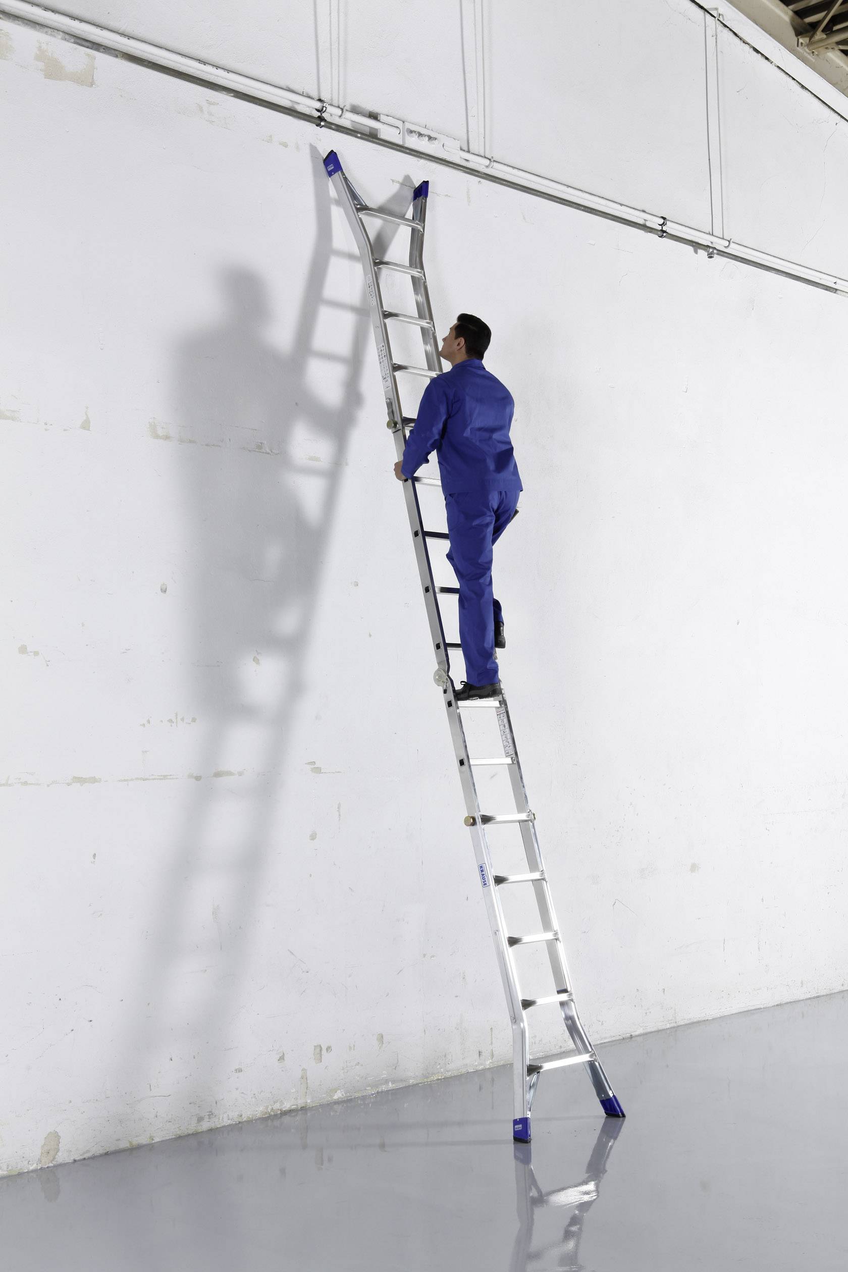 Ein Mann in blauer Arbeitskleidung klettert eine lange Aluminiumleiter, die an einer weißen Wand lehnt, in einem industriellen Raum hoch.
