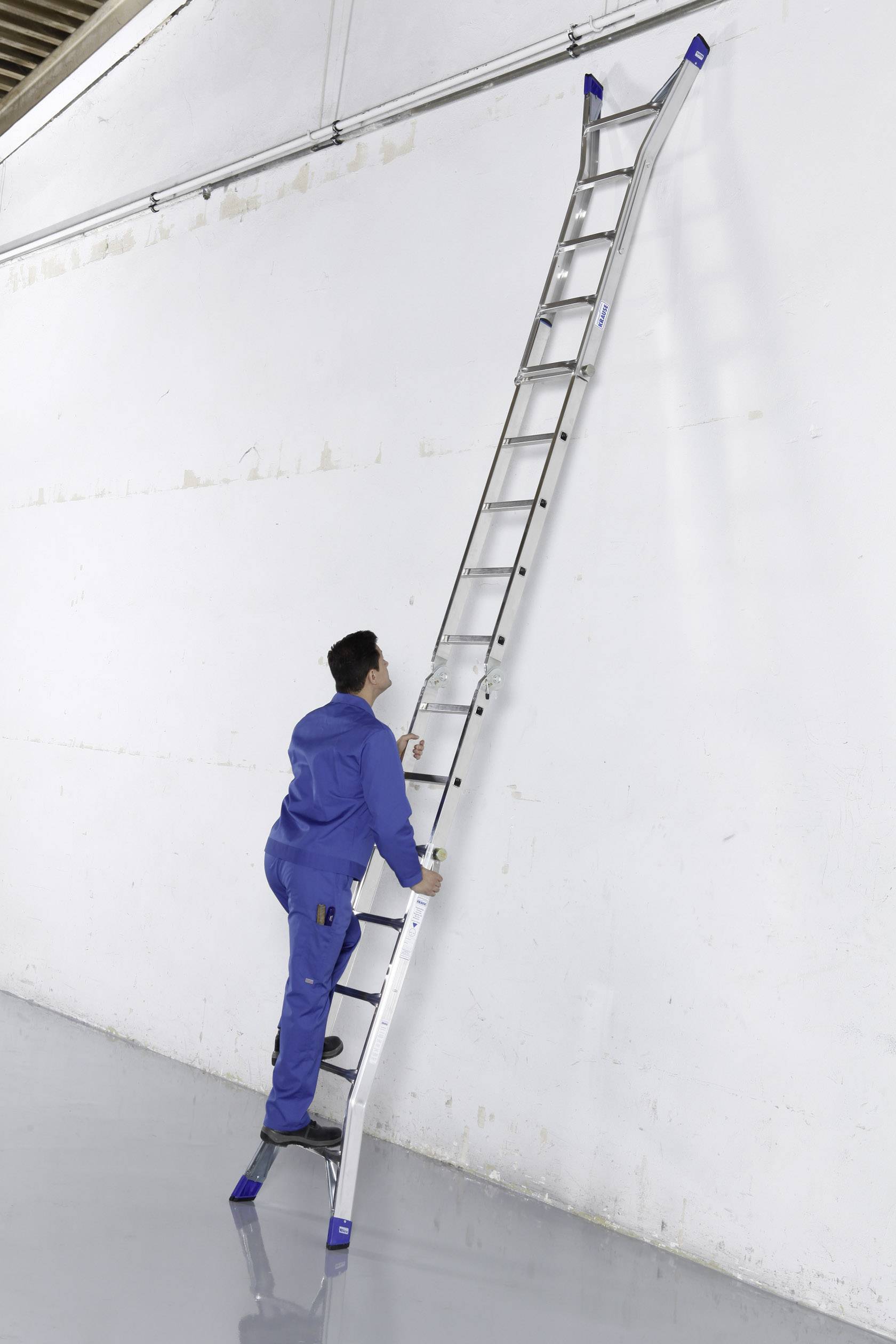 Eine Person in blauer Arbeitskleidung steht auf einer langen, an einer hohen weißen Wand lehnenden Aluleiter.