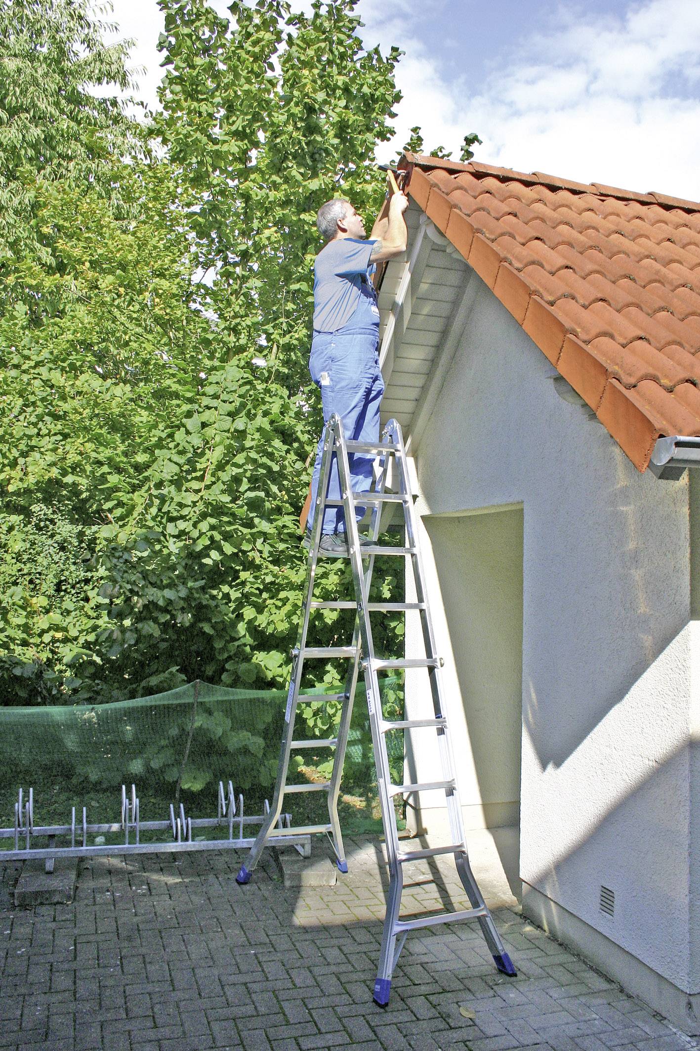 Person steht auf einer hohen Leiter und repariert das Dach eines Hauses im Freien bei Tageslicht, umgeben von grünen Bäumen.