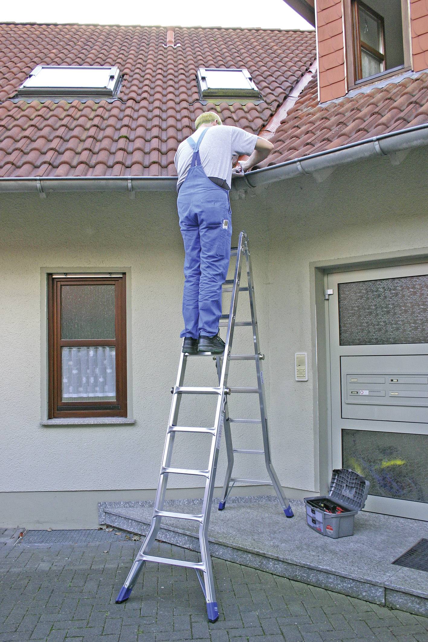 Ein Mann in blauer Arbeitskleidung steht auf einer Leiter und repariert das Dach eines Hauses. Eine Werkzeugbox steht unten.