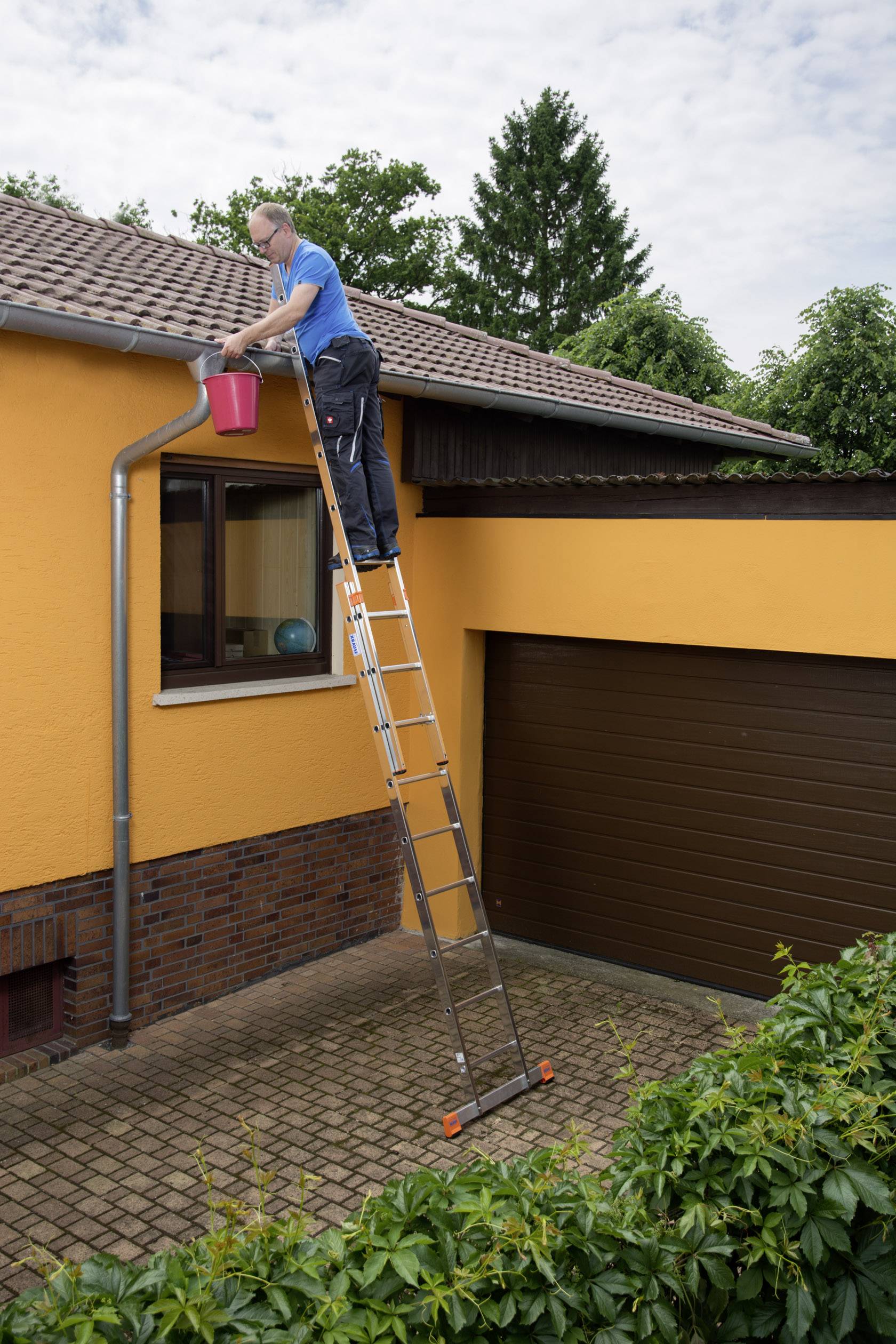 Eine Person steht oben auf einer Leiter und reinigt die Dachrinne eines Hauses mit einem roten Eimer.