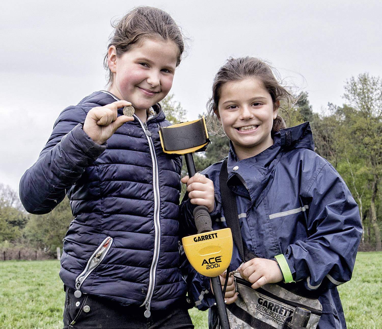 Zwei Kinder stehen fröhlich auf einer Wiese, halten einen Metalldetektor und zeigen stolz einen kleinen Fund in die Kamera.