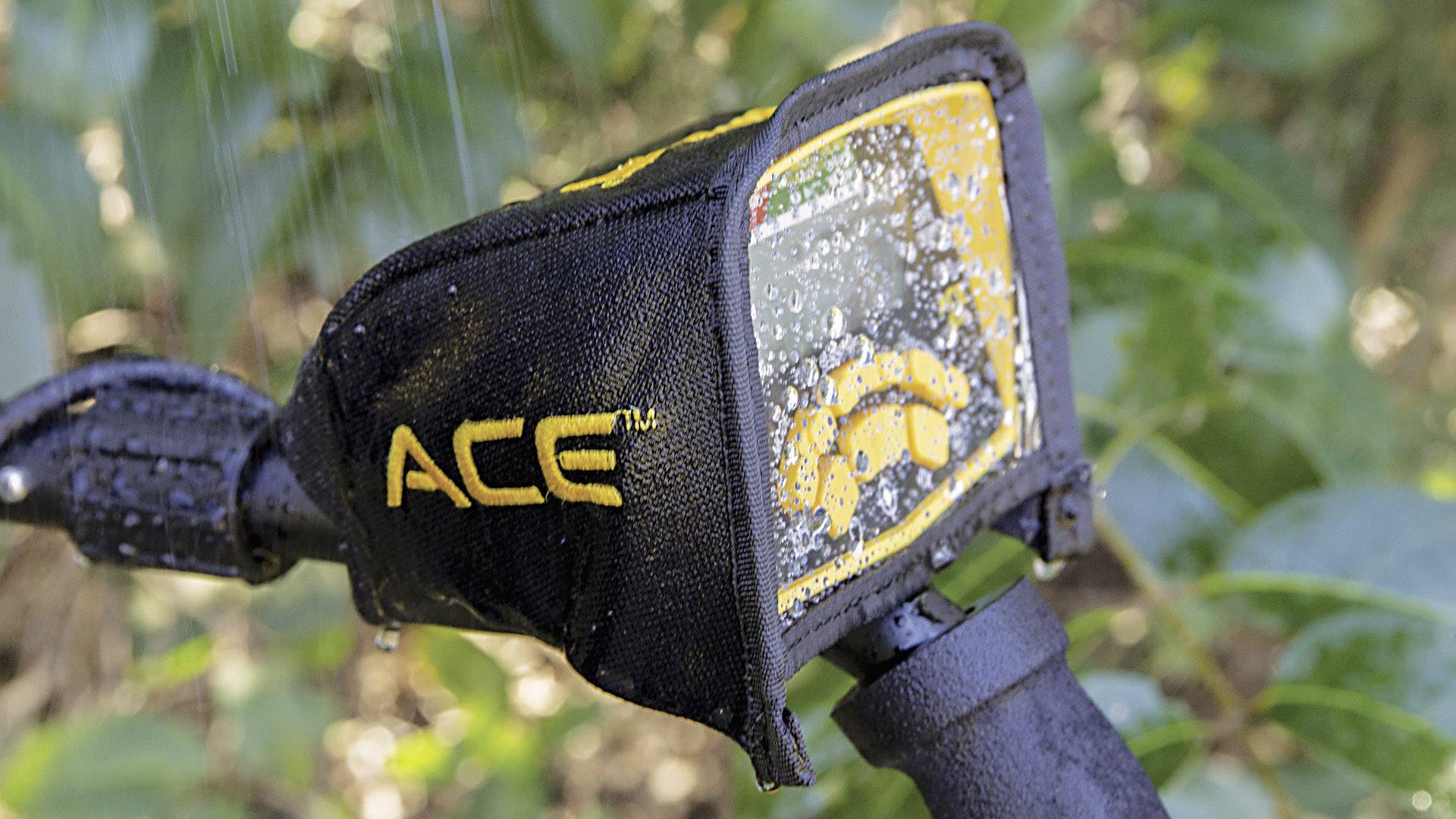 Ein Metalldetektor mit Display wird durchnässt von Wasser. Auf dem Display sind Symbole und eine Skala sichtbar. Im Hintergrund Pflanzen.