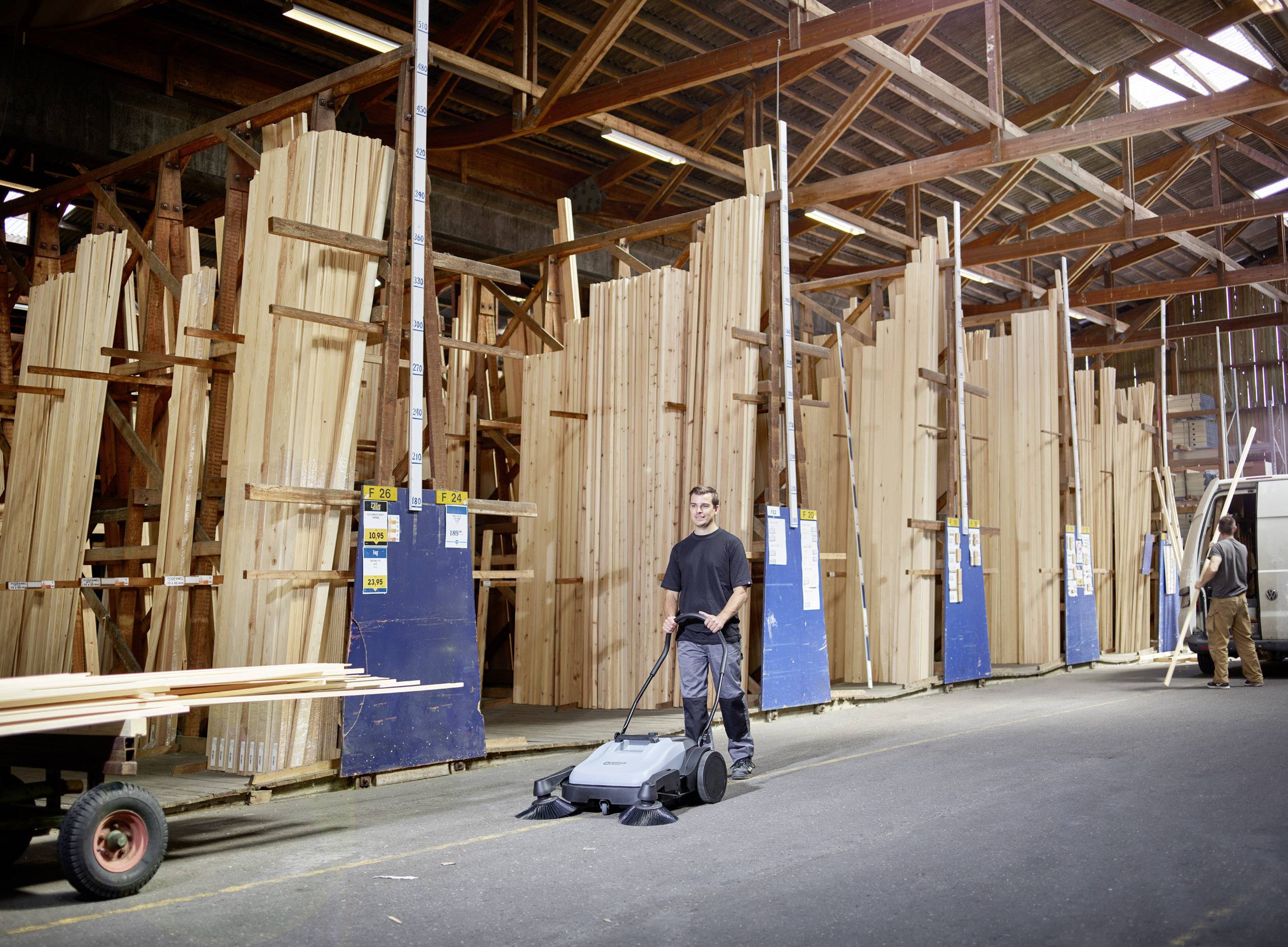 Eine Person kehrt den Boden einer großen Holzbearbeitungswerkstatt. Im Hintergrund stehen hohe Stapel aus rohen Holzbrettern.