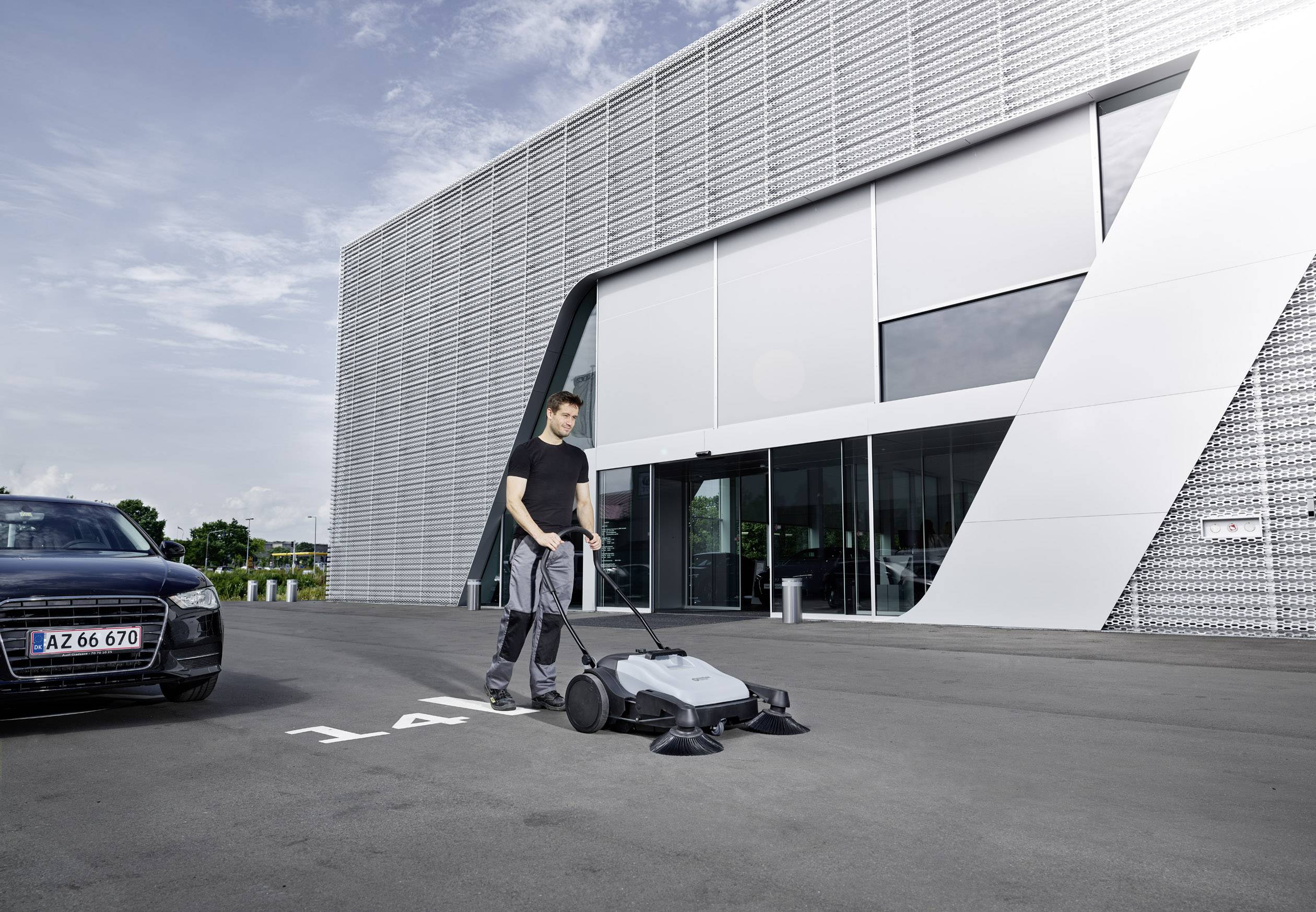 Ein Mann nutzt einen mechanischen Bodenreiniger auf einem Parkplatz vor einem modernen Gebäude bei klarem Wetter.