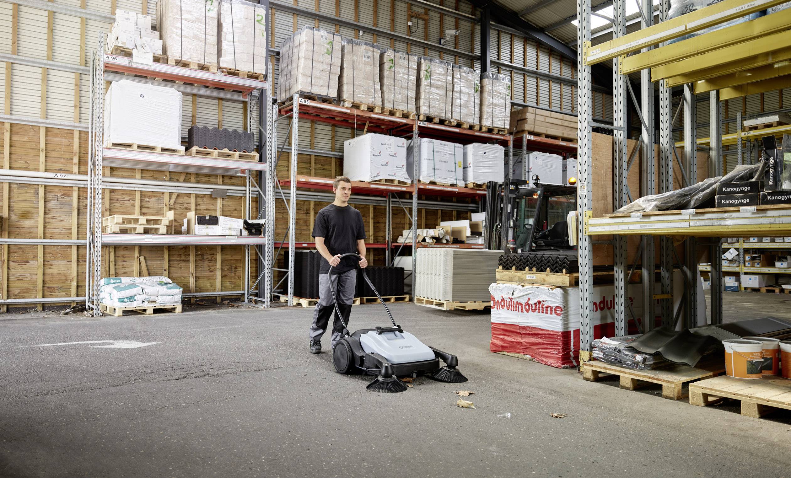 Ein Mann schiebt einen industriellen Kehrmaschine durch ein Lager mit hohen Regalen, auf denen Kisten und Materialien gelagert sind.