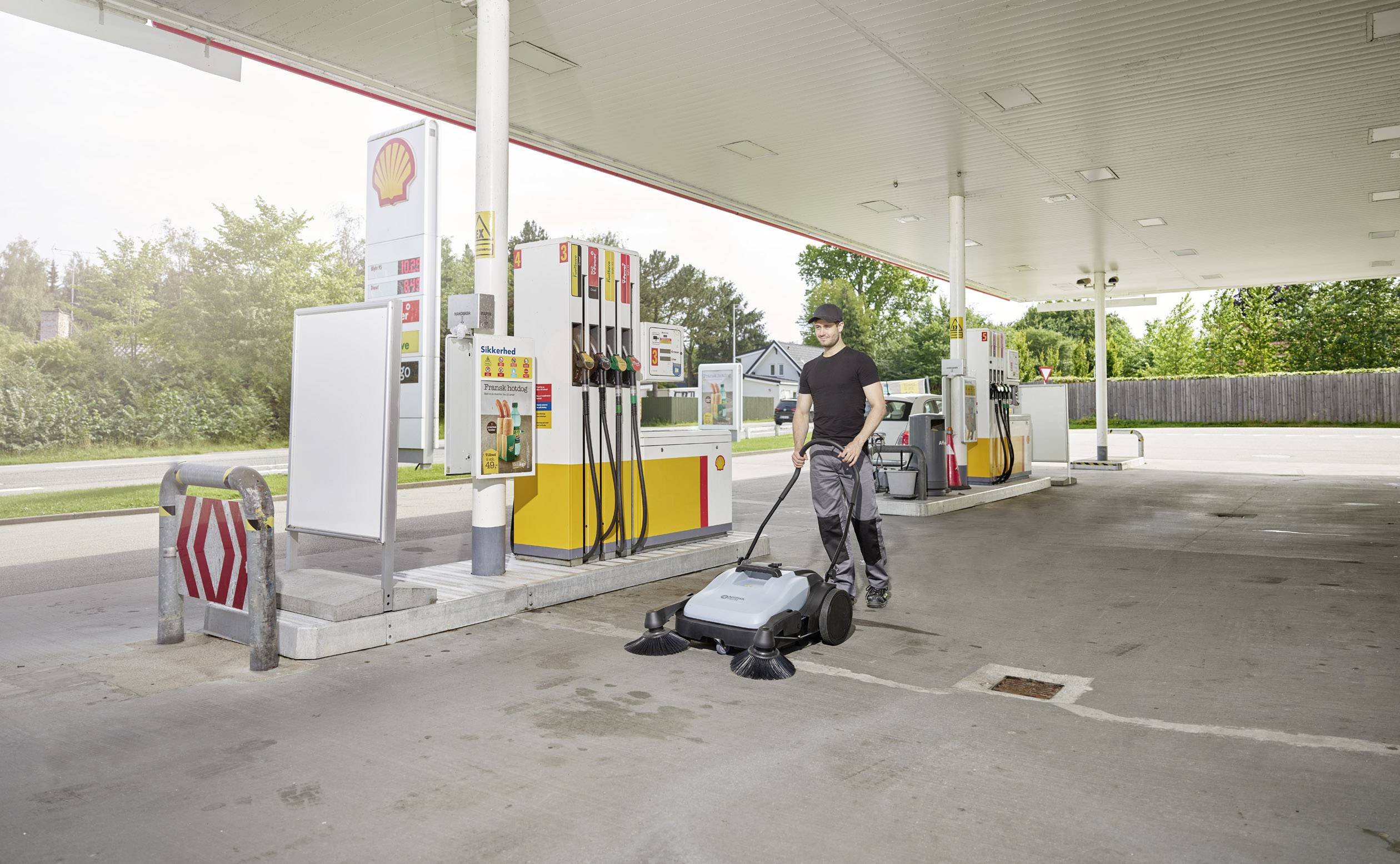 Eine Person reinigt den Boden einer Tankstelle mit einer Kehrmaschine. Im Hintergrund sind Zapfsäulen und Bäume sichtbar.